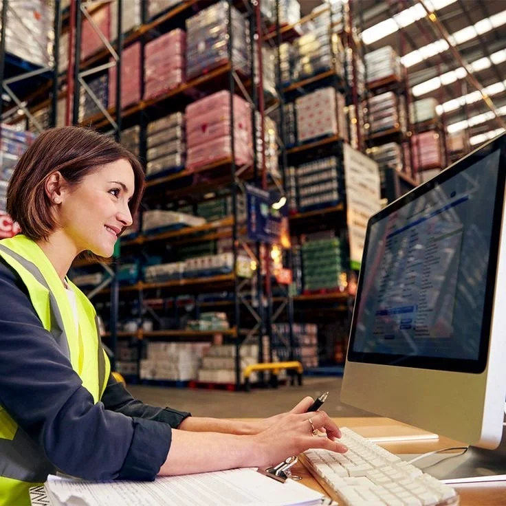 A woman working at a desk in a warehouse, looking at a computer screen, with shelves of stored boxes in the background.