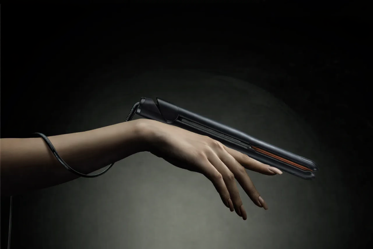 A human hand with manicured nails holding an electric hair straightener against a dark background.