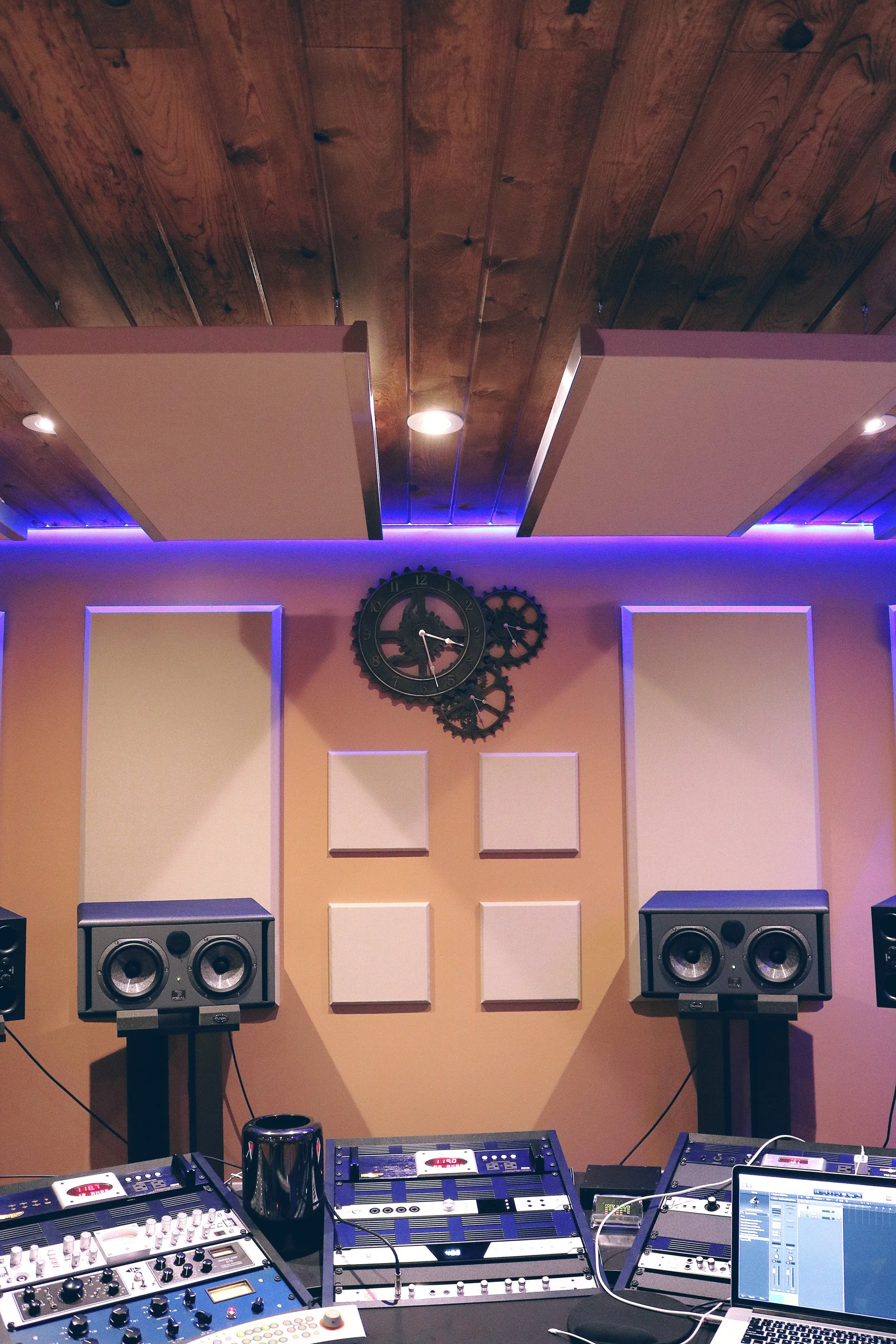 Studio met geluidsisolatie panelen, speakers, en DJ-ontvangst- en bedieningsapparatuur, met een muurklok en decoratieve gearwielen op de muur.
