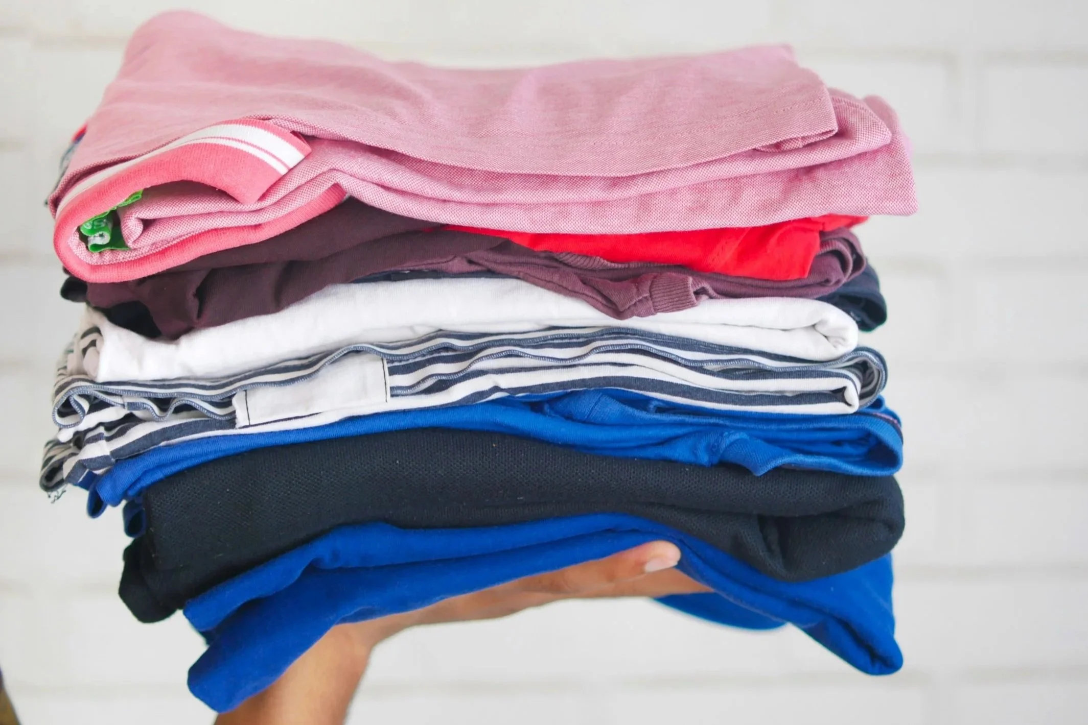 a hand holds a neatly folded stack of clothing in shades of pink, red, white, blue, and navy