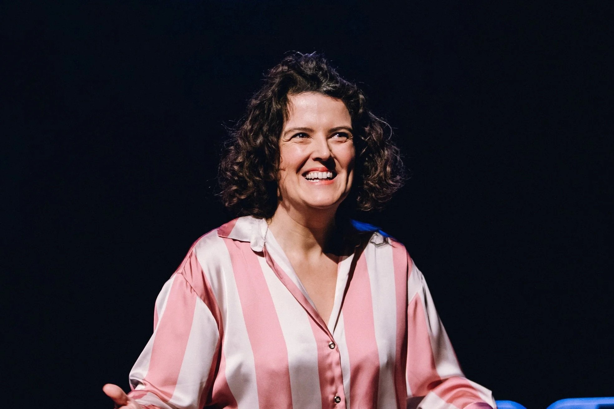 A woman with curly dark hair smiling and wearing a pink and white striped shirt, standing against a dark background.