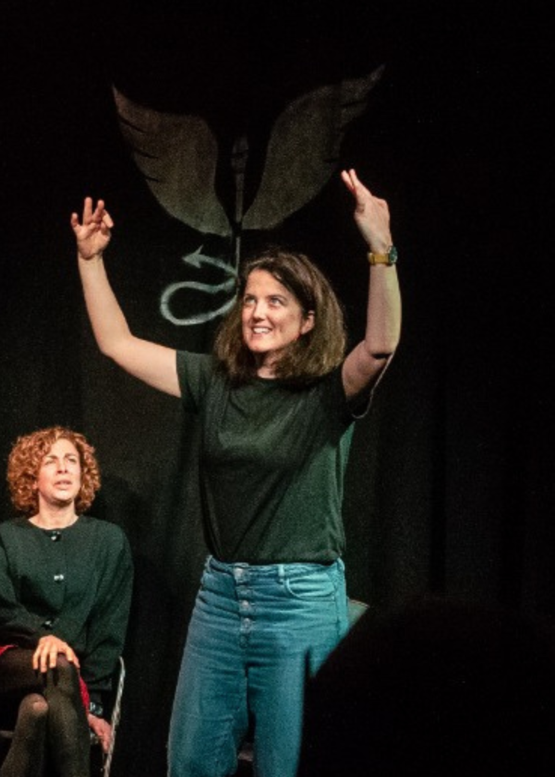 Woman with curly brown hair raising her arms in a 'V' shape on stage, smiling, with a black background and a winged logo above her head. Seated woman with curly red hair is in the foreground, looking to the side.