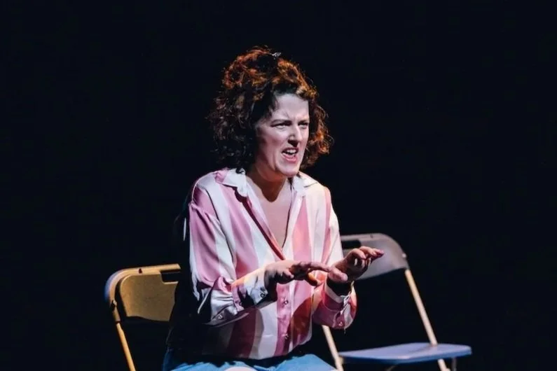 A woman with curly hair wearing a pink and white striped shirt, sitting on a yellow folding chair on a dark stage, making an expressive facial gesture.