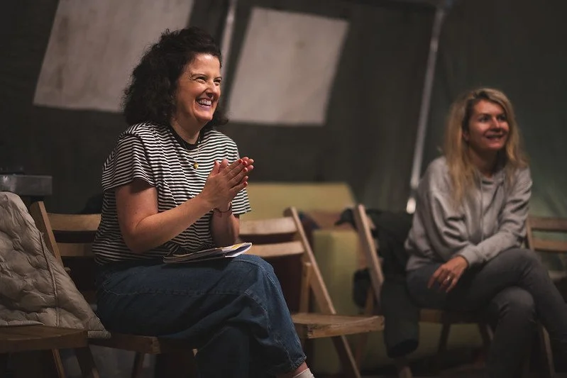 Two women sitting on wooden chairs in a dimly lit room, smiling and laughing, one with curly hair wearing a striped shirt and the other with straight blonde hair in a gray hoodie.