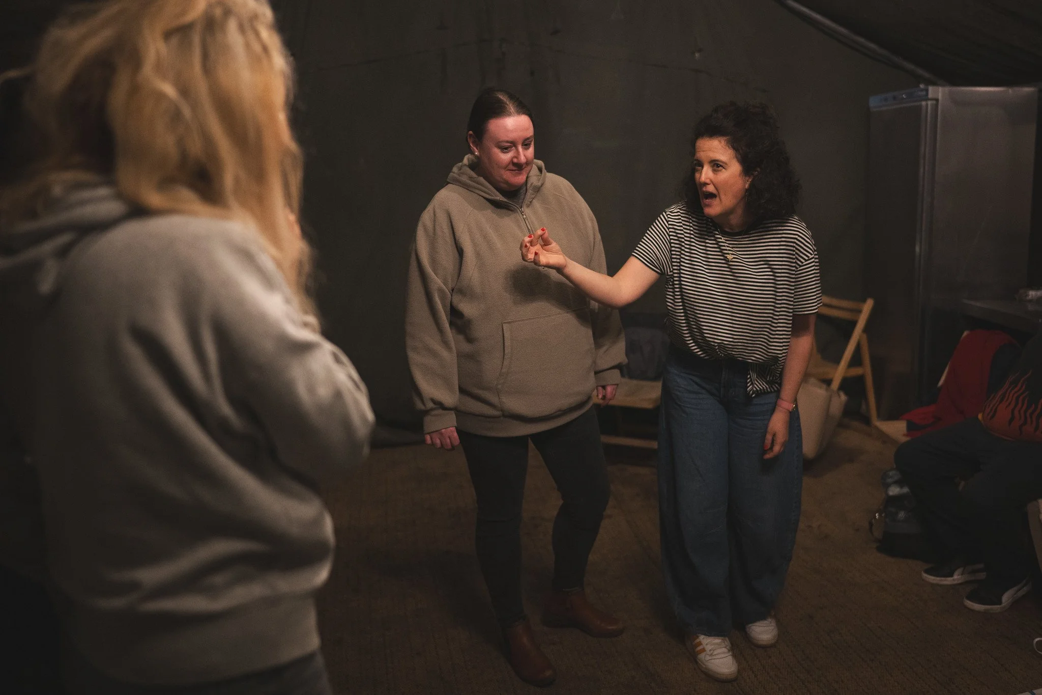 Four women engaged in conversation in a cozy, dimly lit room with dark walls and wooden furniture. One woman is speaking passionately, while others listen attentively.