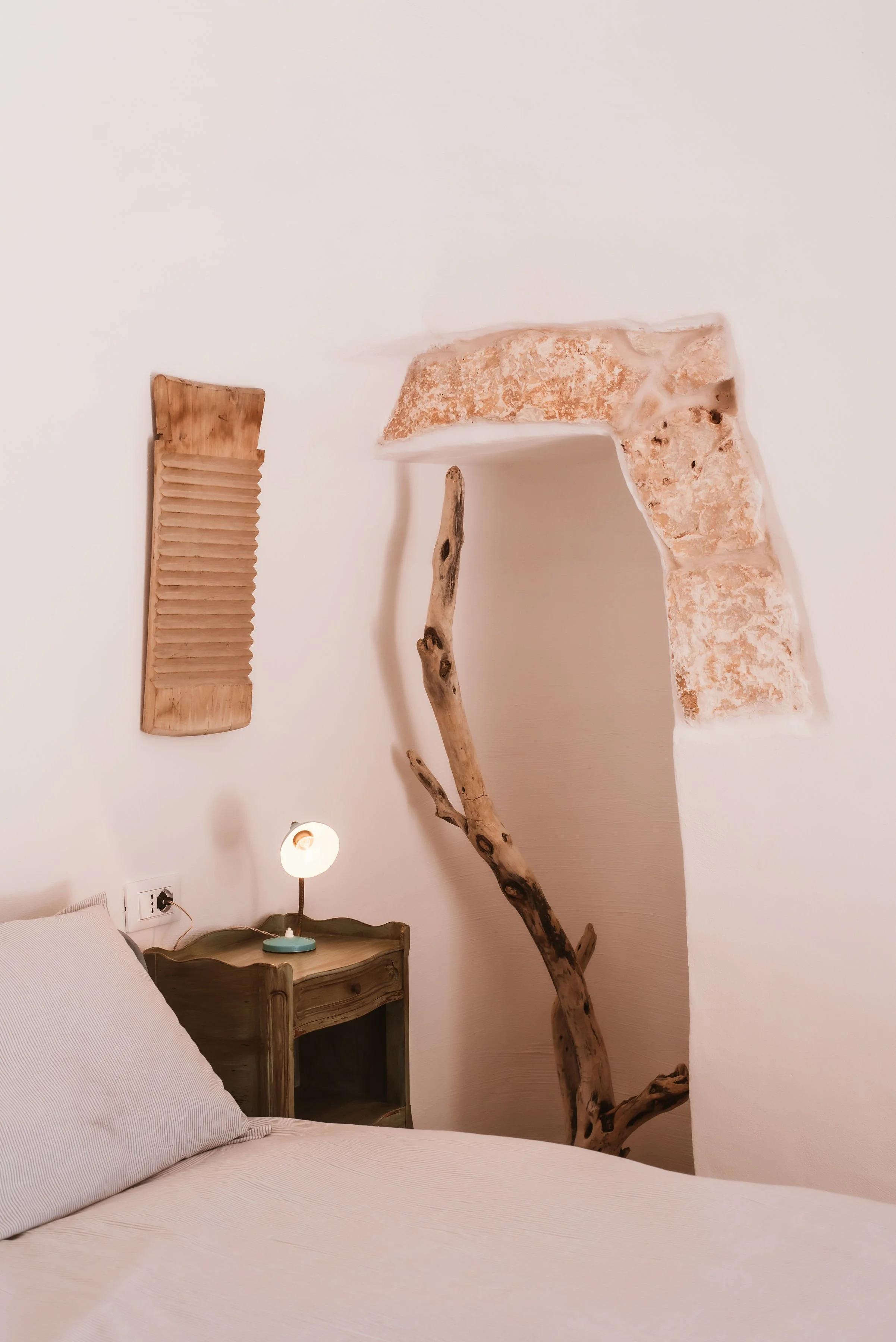 A bedroom with a white bed, a small wooden nightstand with a lamp, a rustic stick leaning against the wall, a wooden washboard hanging on the wall, and exposed brickwork around a small archway in the wall.