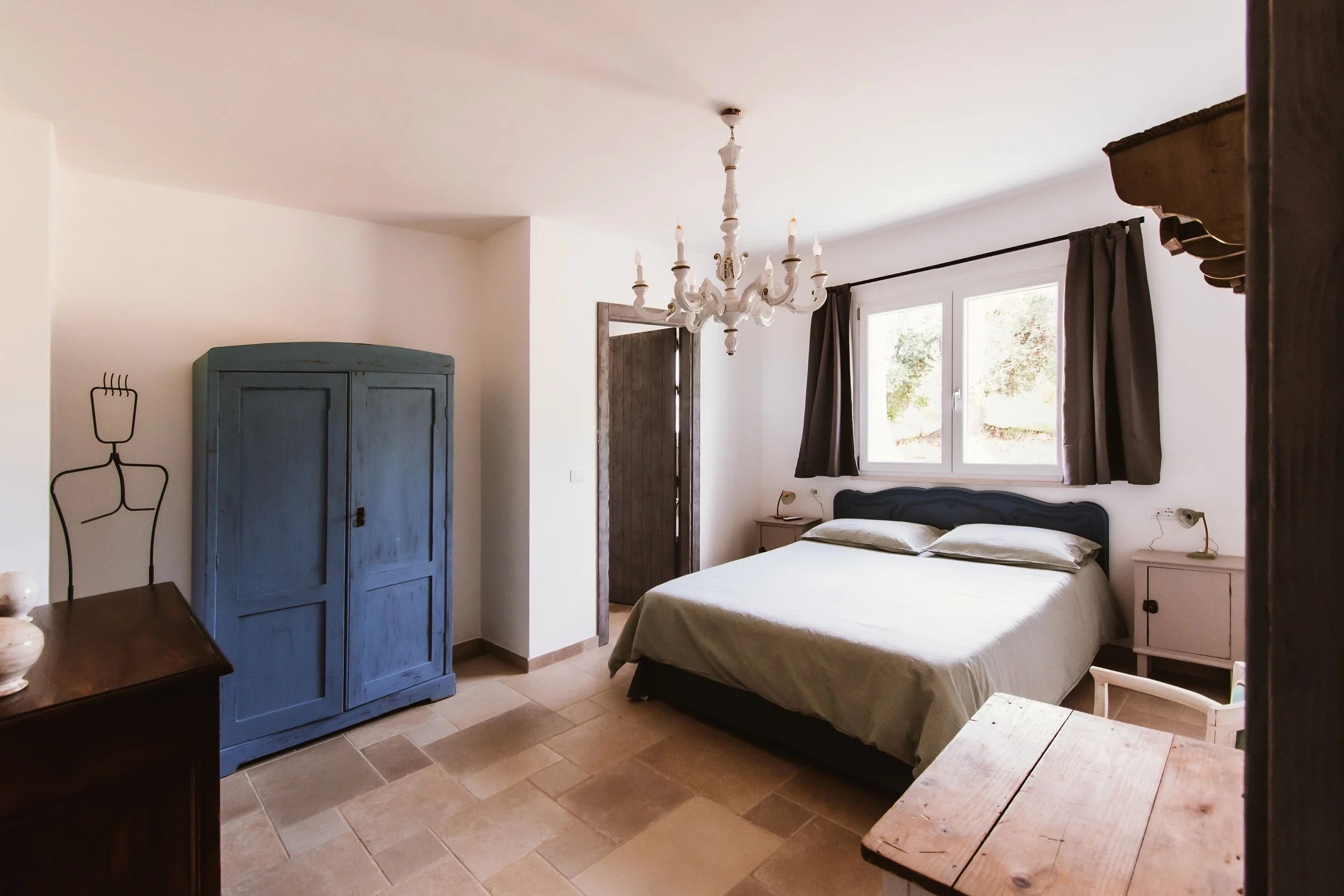 A bedroom with a bed, nightstands, a blue wardrobe, a chandelier, and a window with dark curtains.