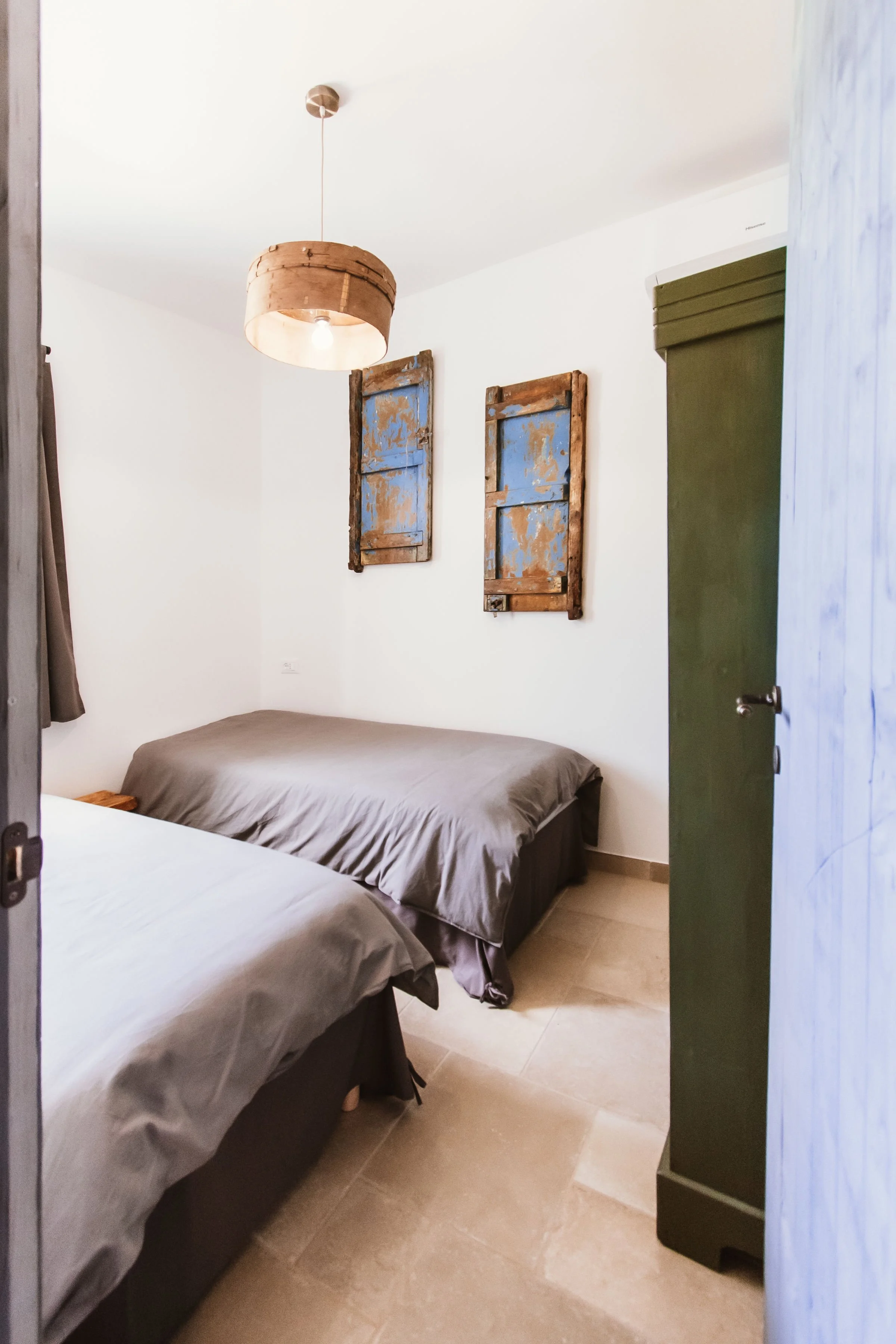 A bedroom with two beds, each with gray bedding. The wall has two rustic, blue and brown wooden window frames as decoration. There is a brown hanging light fixture and a green wardrobe on the right side. The room has beige tiled flooring and natural