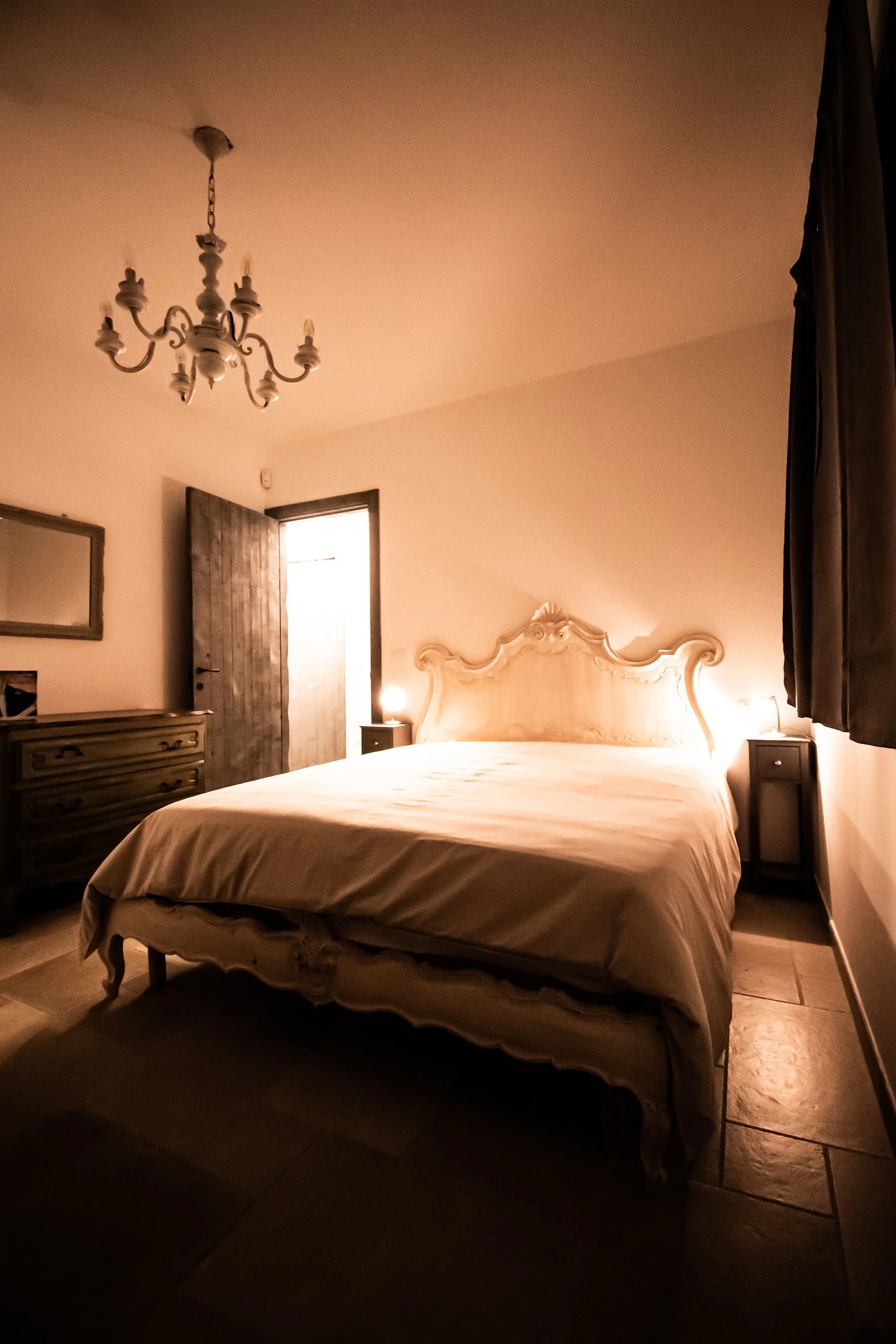 A cozy bedroom with a vintage style, featuring a wooden bed with an ornate headboard, two nightstands with lamps, a dresser, a mirror, a wooden door, and a chandelier hanging from the ceiling.
