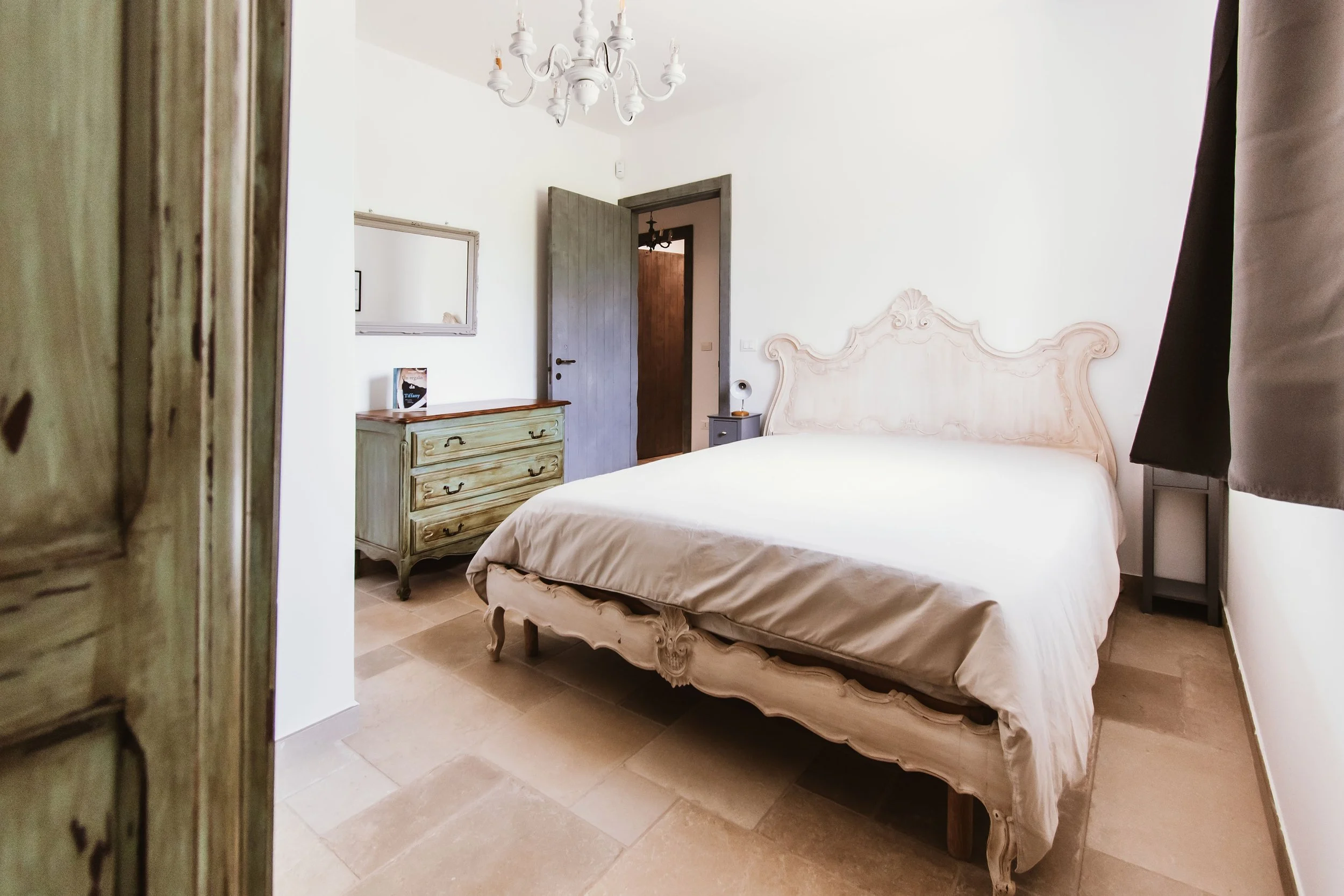 A bedroom with a vintage style, featuring a large bed with an ornate headboard, a greenish dresser, a large mirror, and a small dark side table with a lamp. The room has white walls, a chandelier, and a partially open door leading to another room.