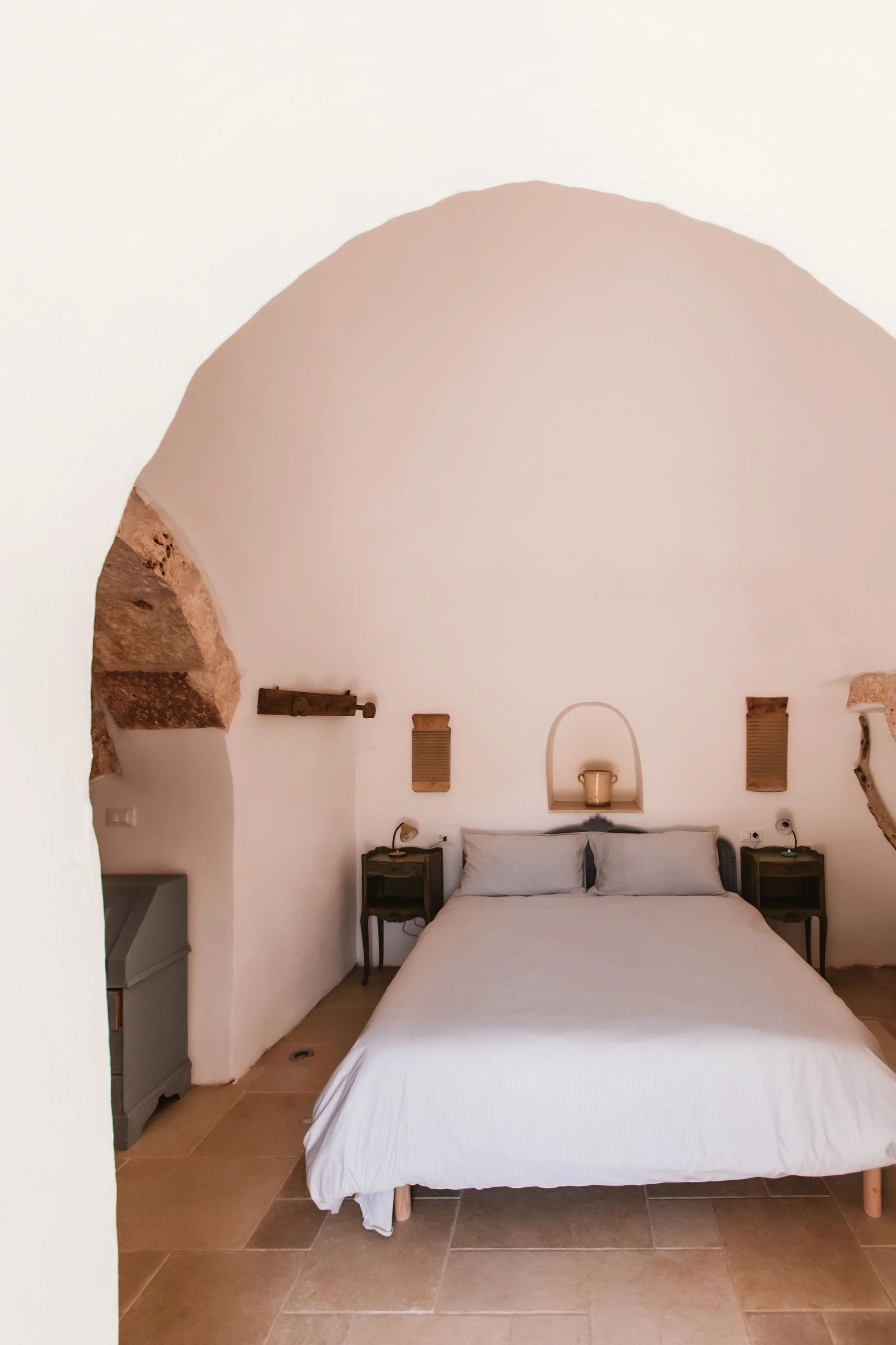 A minimalist bedroom with a white bed, two bedside tables, and rustic wall decorations in a room with a white curved ceiling and stone accents.