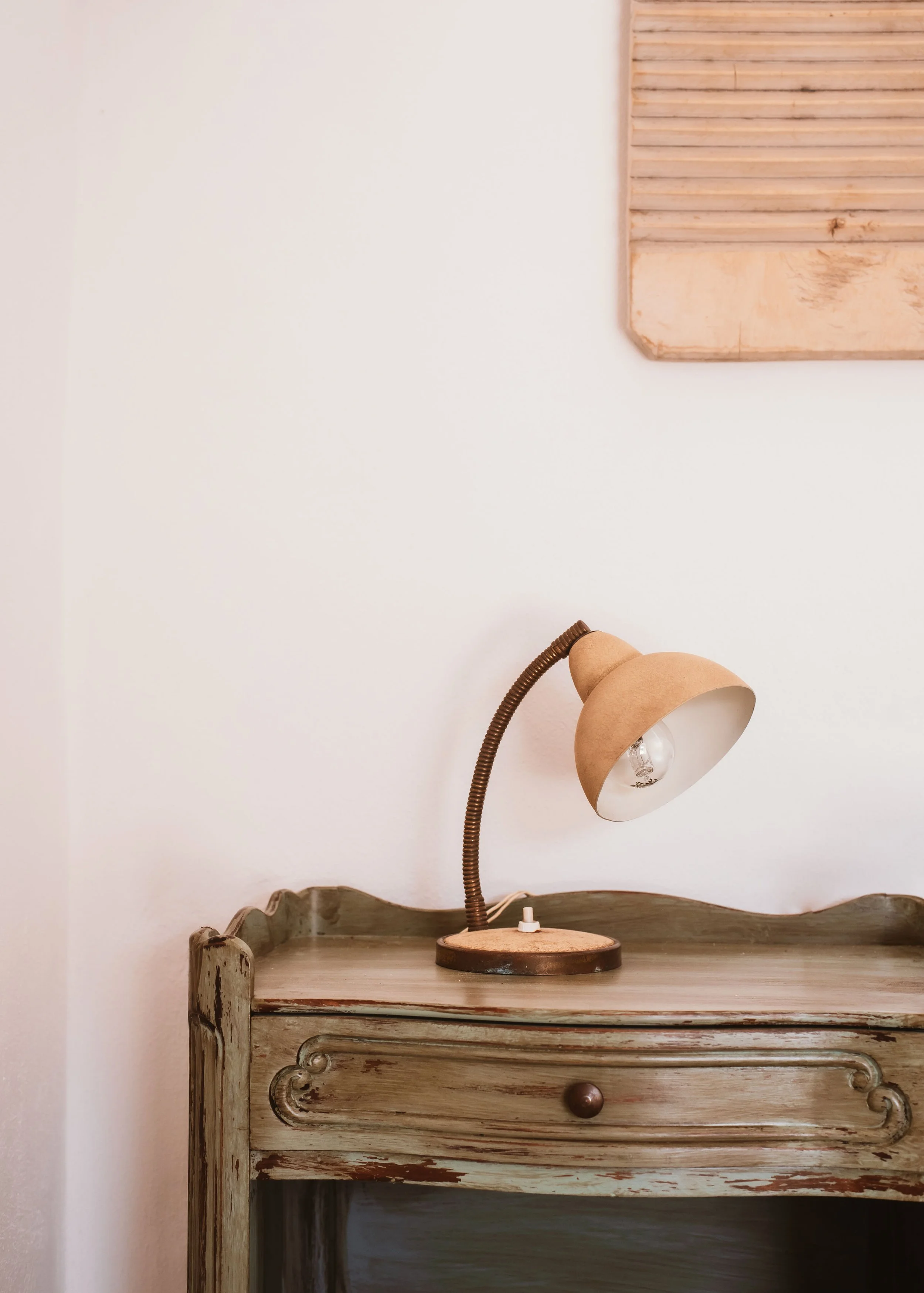 A vintage wooden nightstand with a distressed finish, a curved top, and a single drawer with a round knob. On top of the nightstand is a beige desk lamp with a flexible neck and a clear lightbulb. Part of a framed wooden wall decoration is visible on
