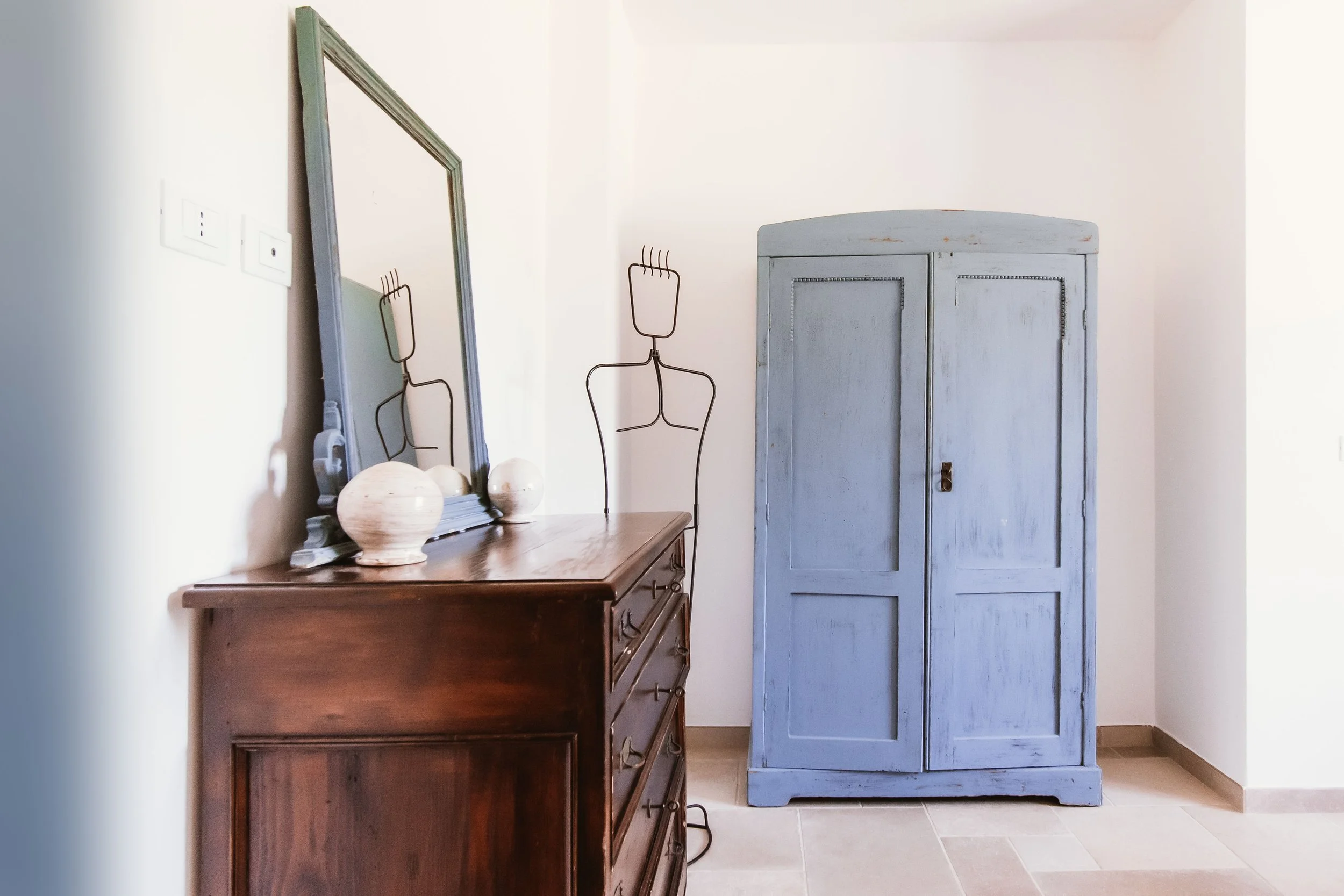 A room with a wooden dresser with a mirror on top, decorated with ceramic vases, next to a blue-painted wooden wardrobe, and two wire sculptures of human figures standing beside the dresser.