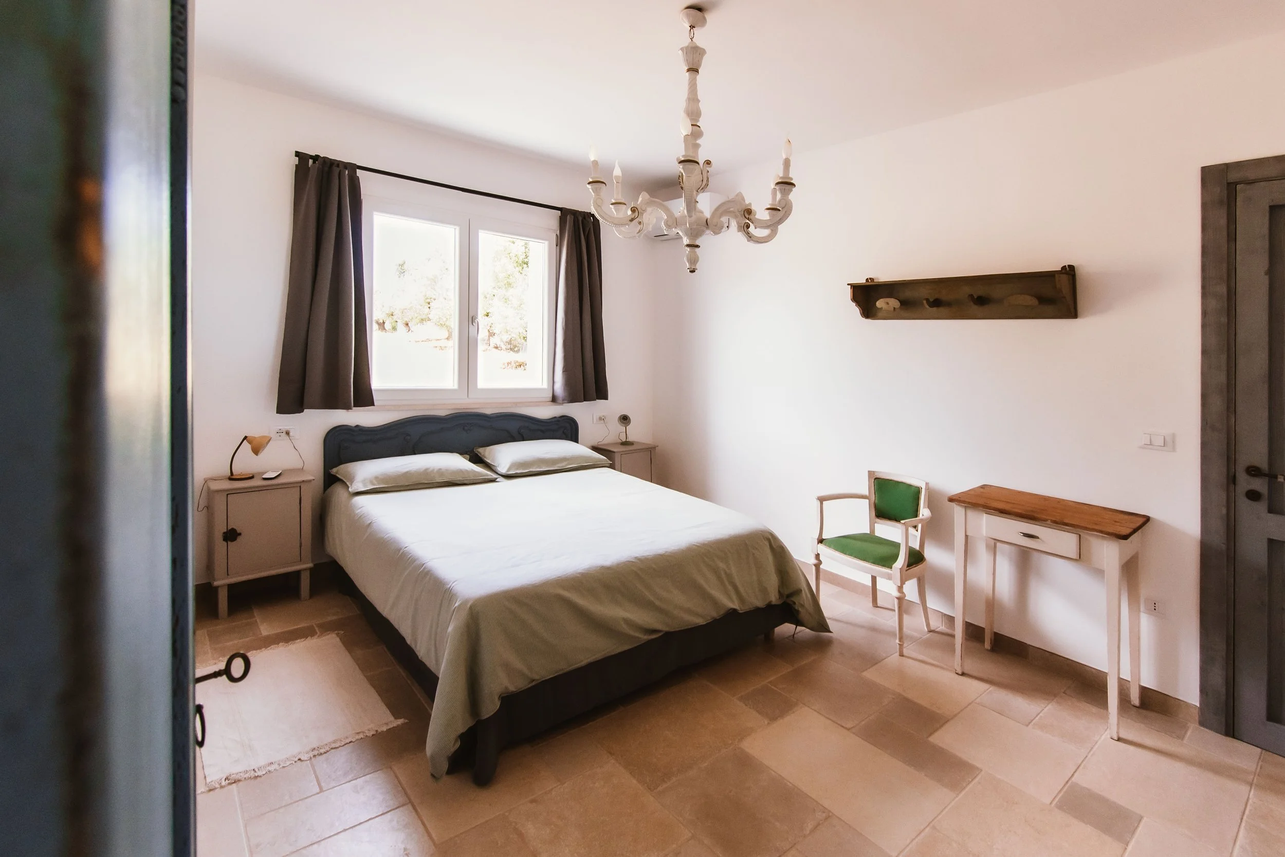 A bedroom with a bed, nightstands, a chair, a desk, and a chandelier.