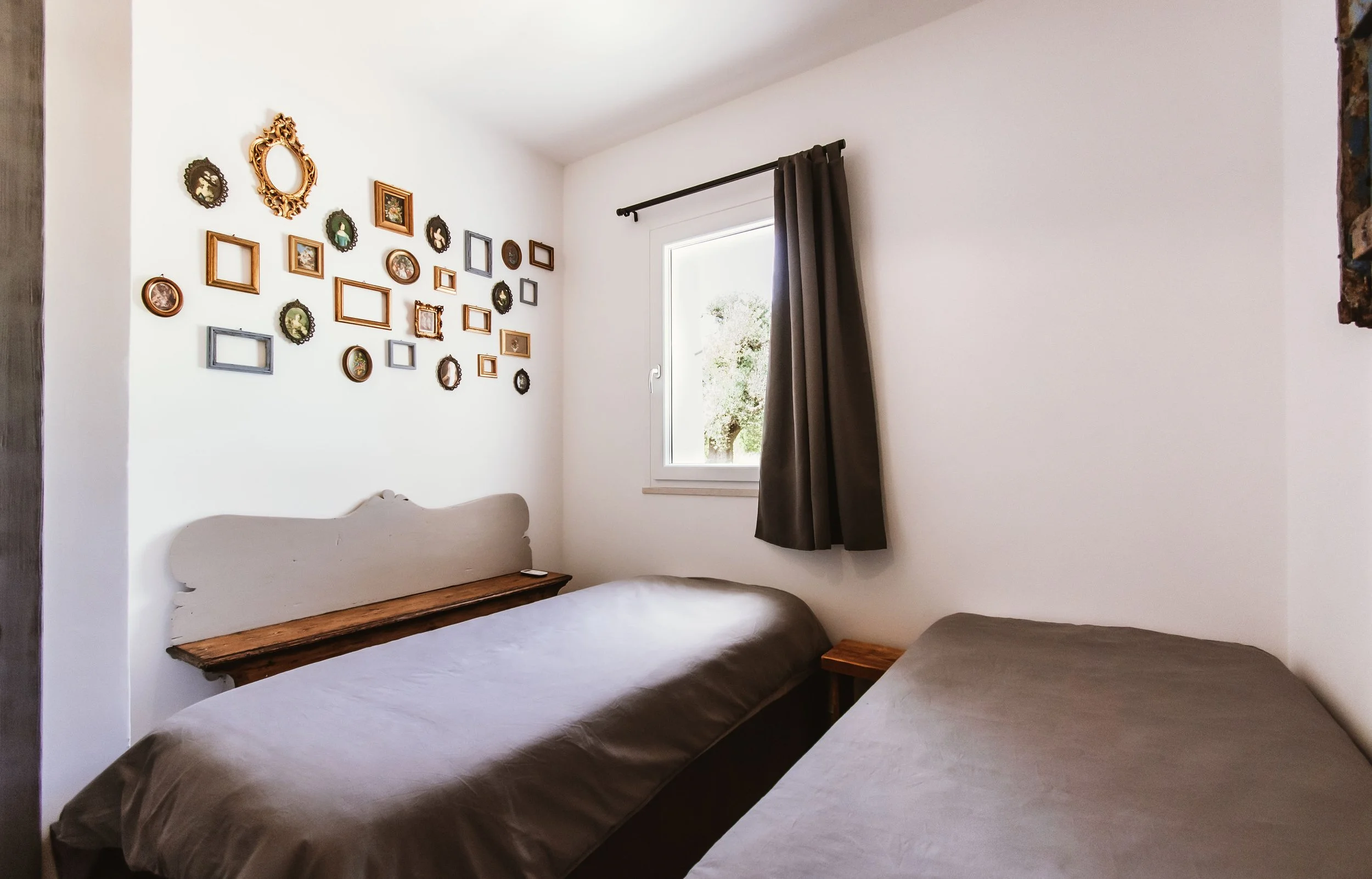 A small bedroom with two single beds covered in brown blankets, a window with dark curtains, a gallery wall with various picture frames, and a wooden nightstand.