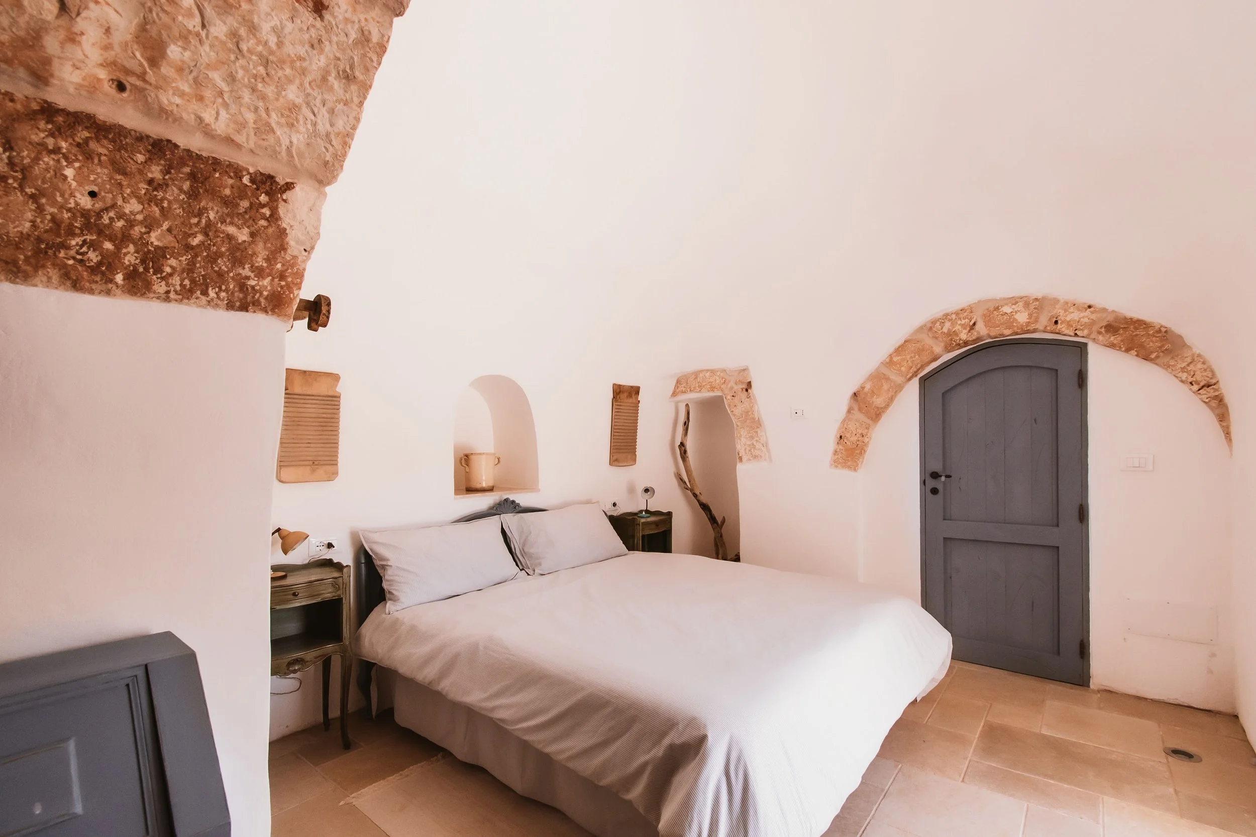 A bedroom with rustic design, white walls, an arched blue door framed with brick, bed with beige bedding, and small wall niches with decor.