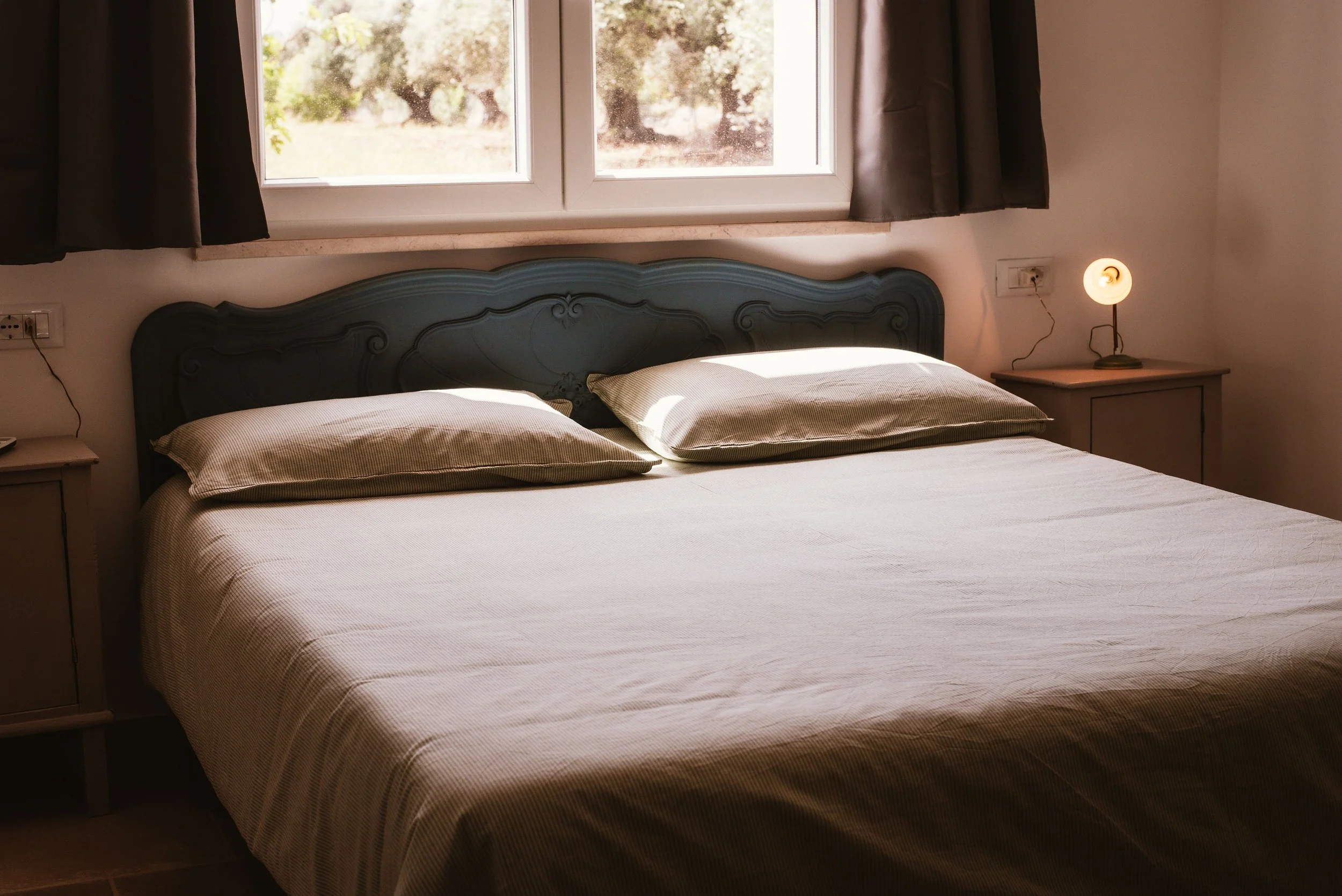 A neatly made double bed with beige pillows and a beige bedspread, located in a cozy bedroom with a window above the headboard showing outdoor trees, flanked by dark curtains. There is a nightstand on each side of the bed, with a small lamp on the ri