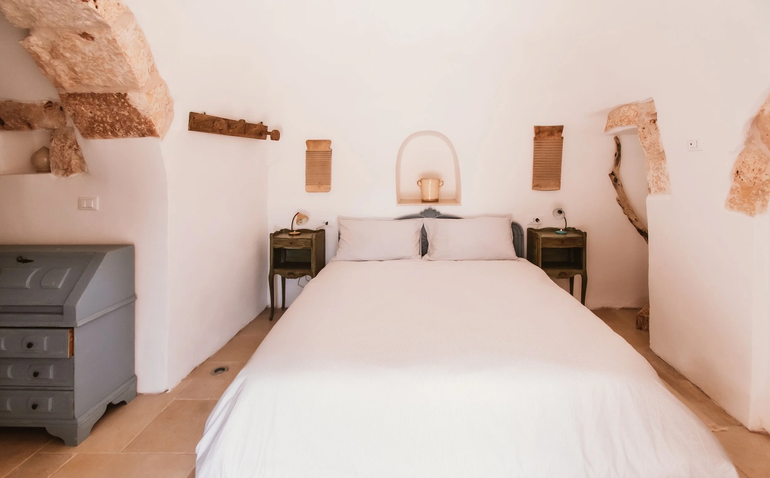 A minimalist bedroom with white walls, a large bed with white bedding, two dark wood nightstands, and rustic decor such as wooden shelves and a decorative arch with a lantern above the bed.