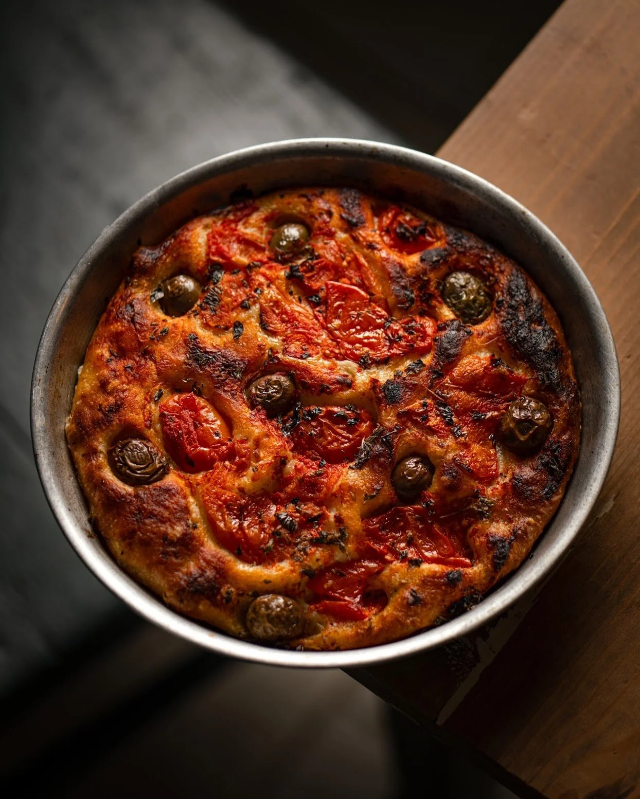 Baked pizza with cherry tomatoes and black olives in a metal pan on a wooden surface.