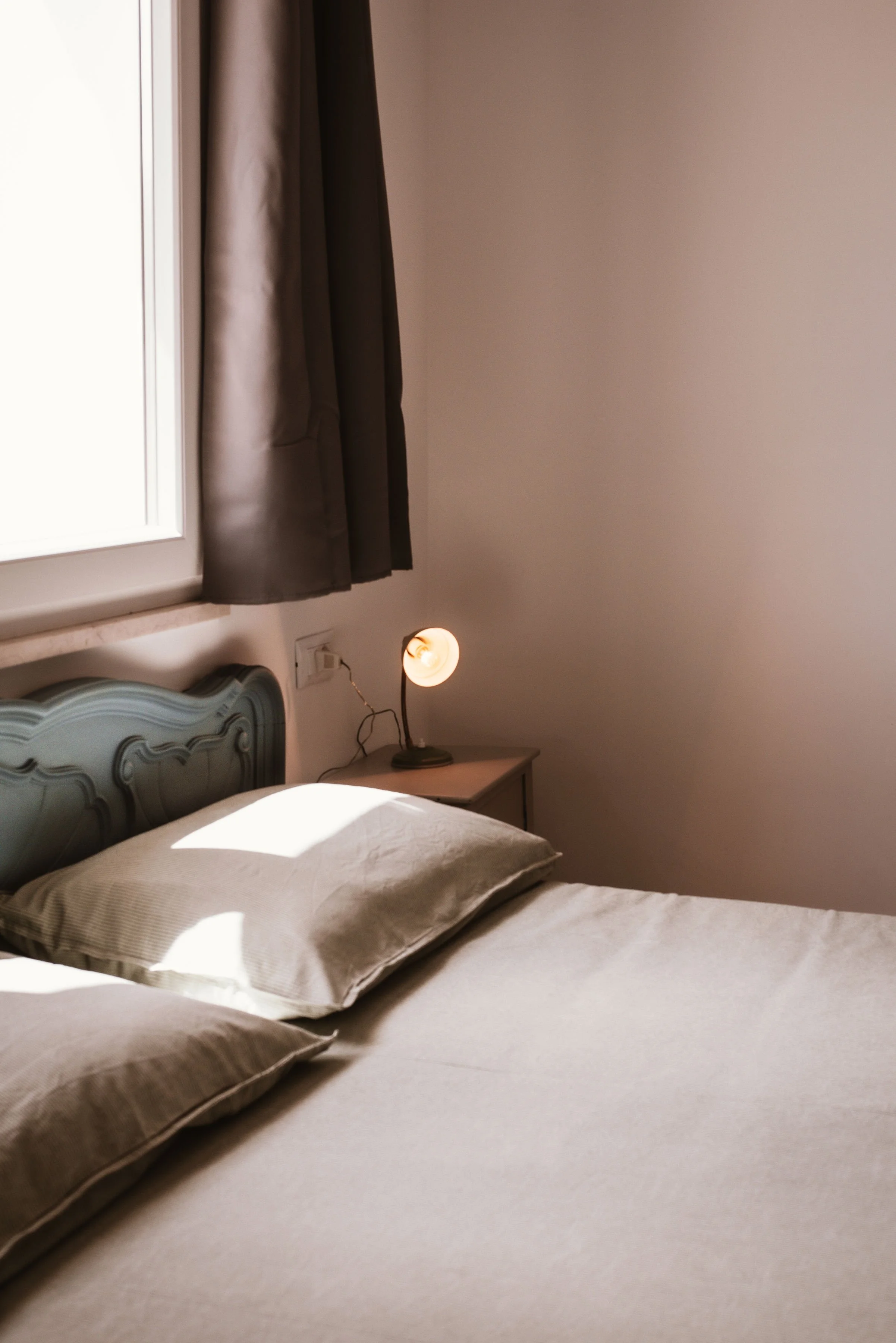 A cozy bedroom with a bed, beige bedding, a headboard, a nightstand, a small lamp, and a window with curtains.