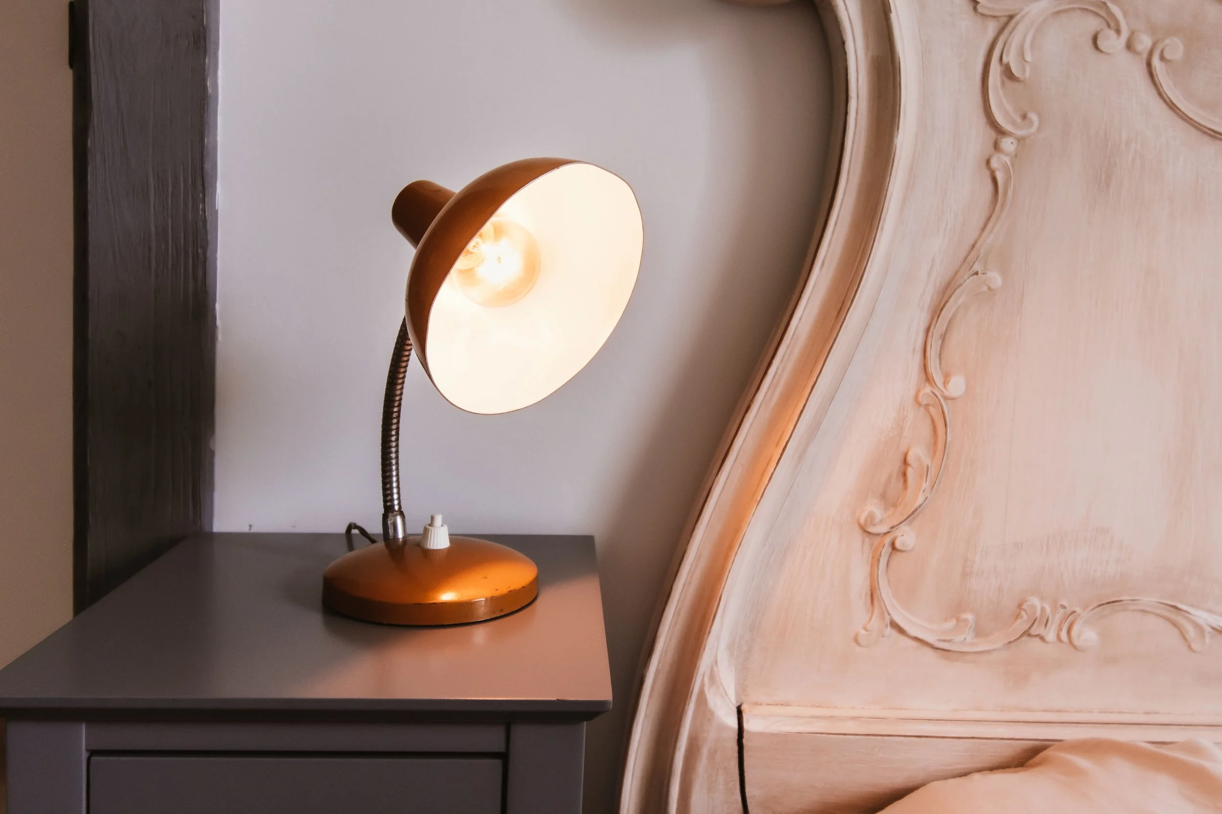 A copper-colored adjustable desk lamp on a gray nightstand next to a carved wooden headboard and a beige pillow.