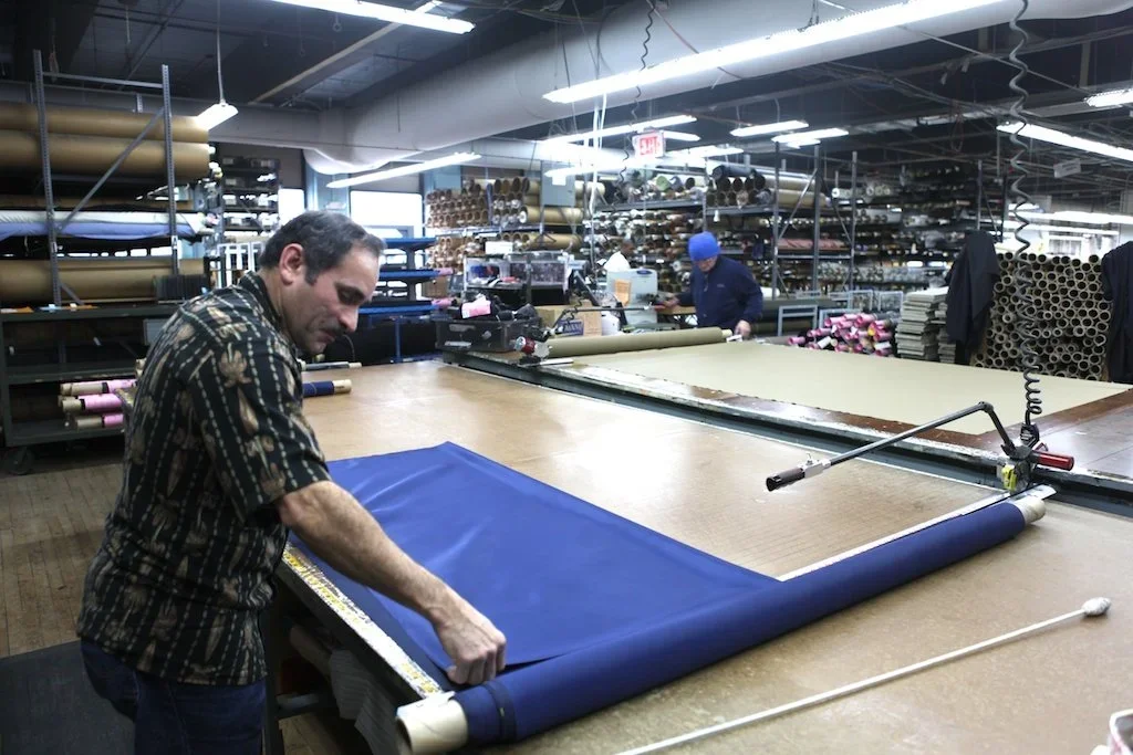 Two workers in a factory working on fabric production; one in the foreground unrolling a large roll of blue fabric and the other in the background handling pink and purple fabric rolls.