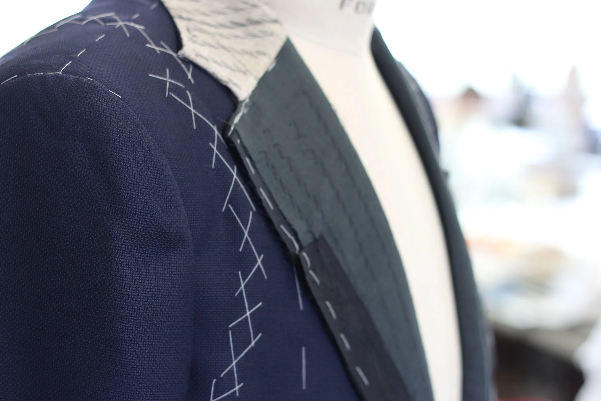 Close-up of a person's shoulder and neck, wearing a navy blazer with white stitch detailing and a white shirt underneath, with a blurred background.