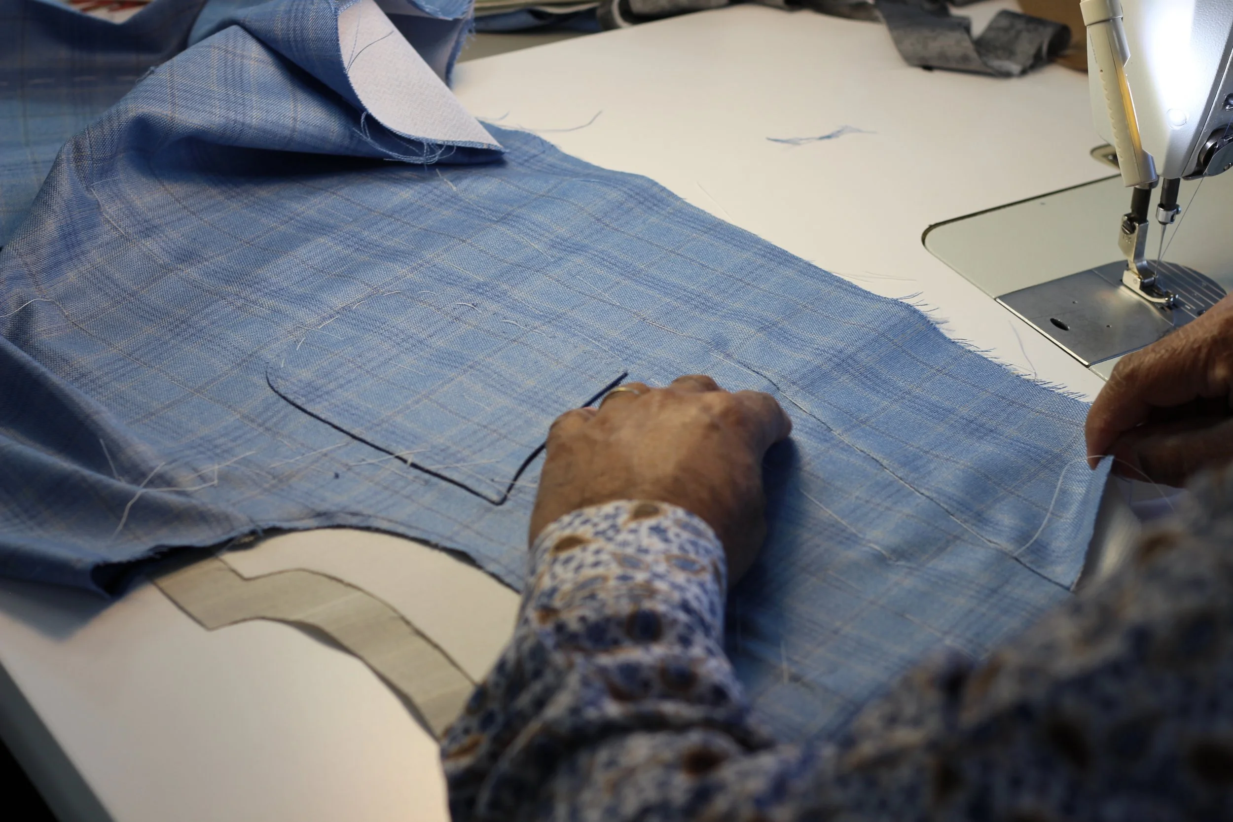 A person sewing a piece of blue fabric on a sewing machine, with another piece of fabric underneath, in a sewing workspace.