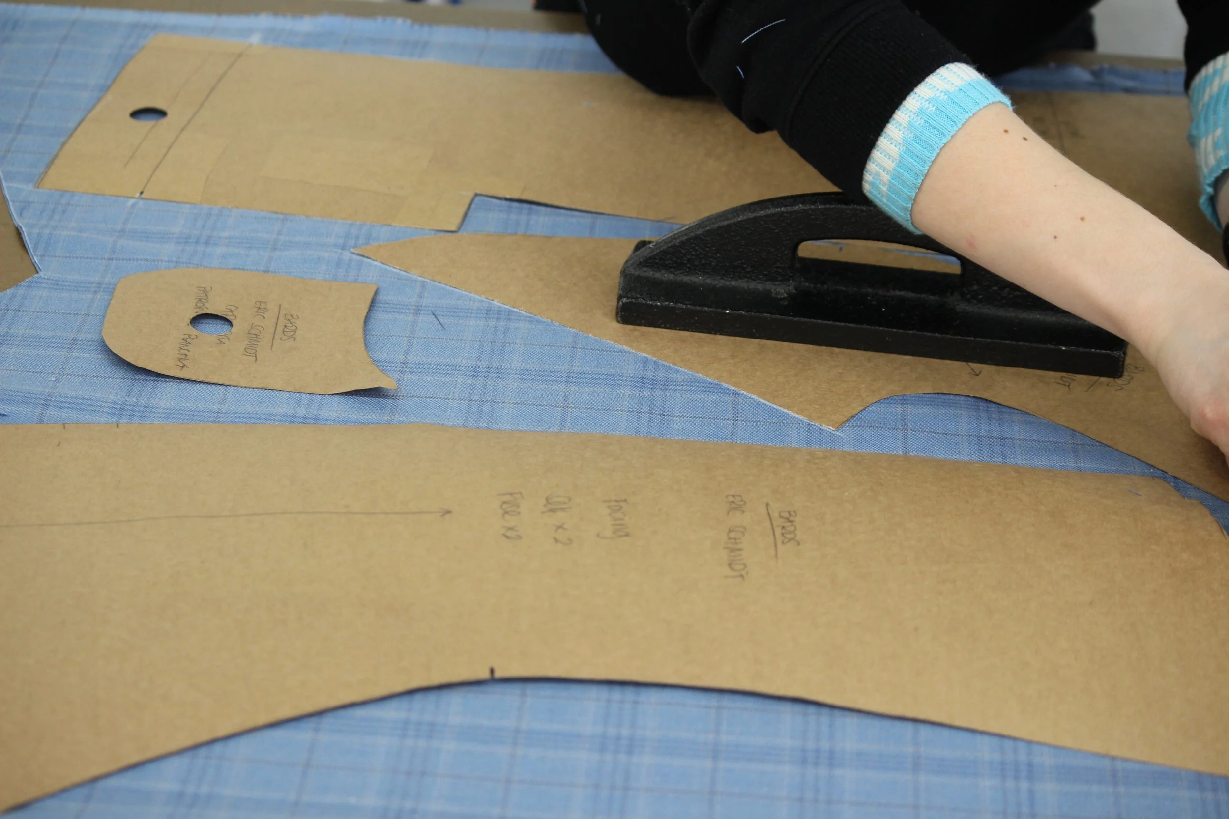 A person is cutting a pattern piece from brown cardboard with a black paper cutter. The pattern piece has some handwritten notes on it, and there are other patterned and cardboard pieces on the table, which is covered in a blue grid-patterned fabric.