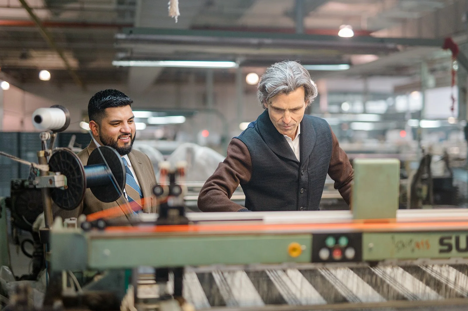 Two men in a factory, one older with gray hair wearing a dark vest, and one younger with dark hair and a beard wearing a suit and tie, working together on a machine.