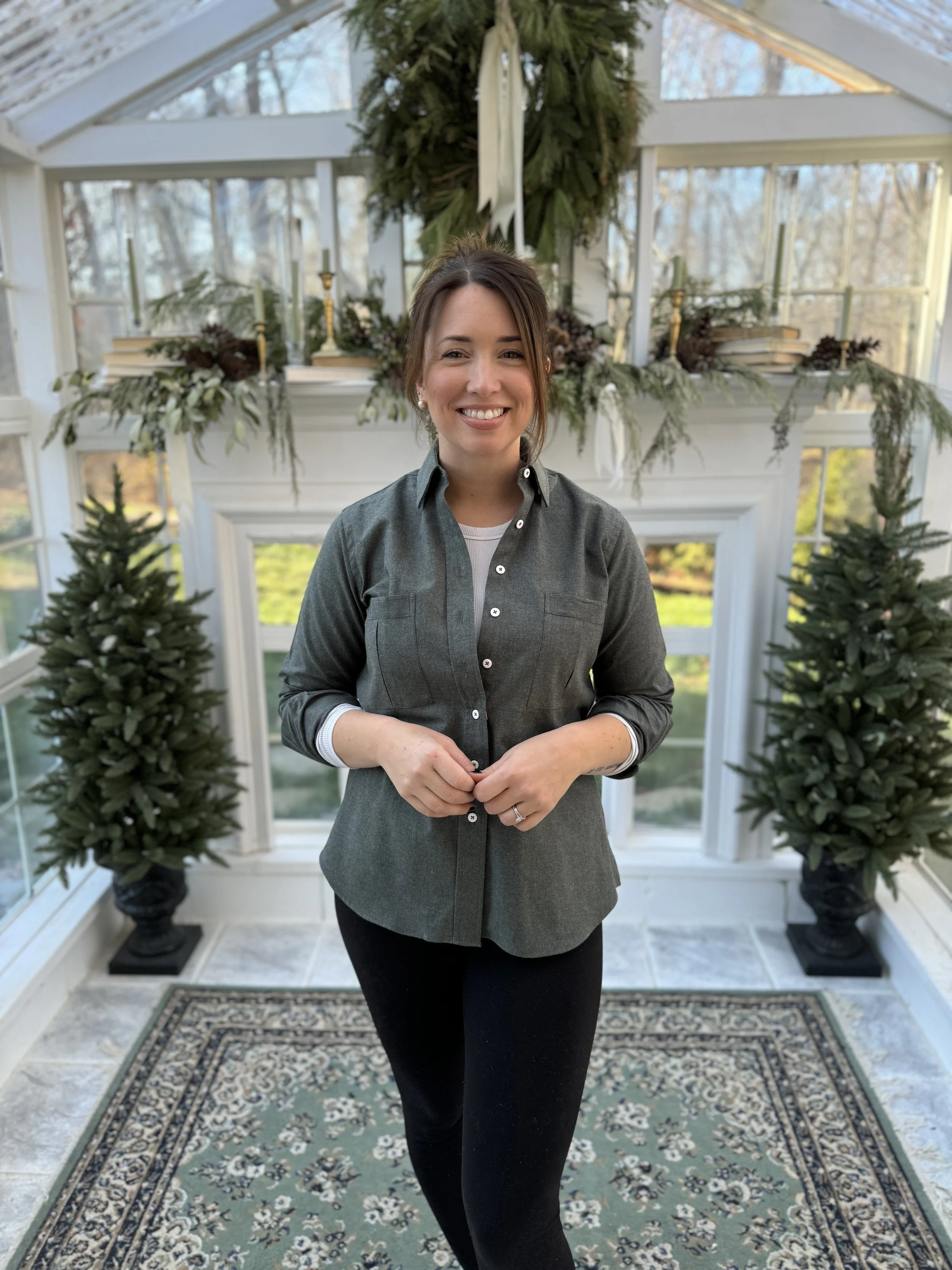 A woman standing indoors in front of a decorated fireplace with Christmas greenery, small potted Christmas trees, and a patterned rug.