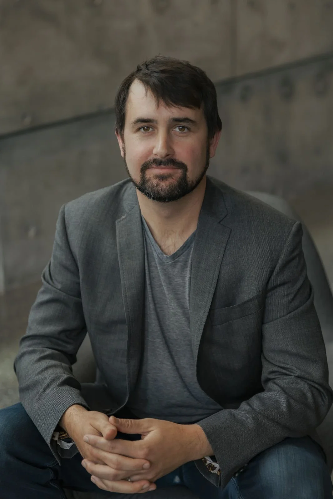Portrait of a man with dark hair, beard, wearing a gray blazer over a gray t-shirt, sitting with hands clasped in front of him, against a dark, textured background.