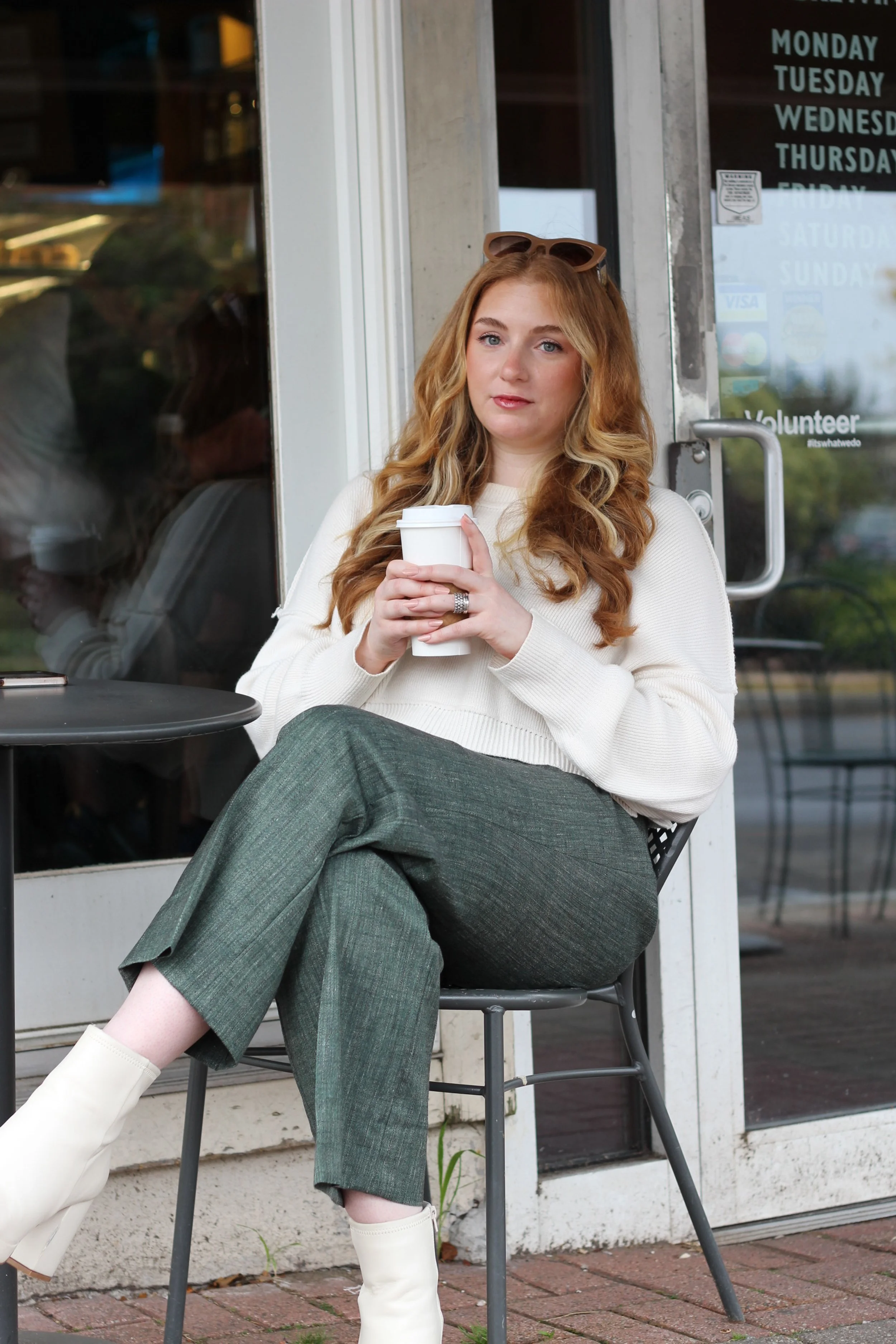 A woman with long blonde hair, wearing a white sweater, green checked pants, and white boots, sitting outside a coffee shop with her legs crossed, holding a disposable coffee cup, looking at the camera.