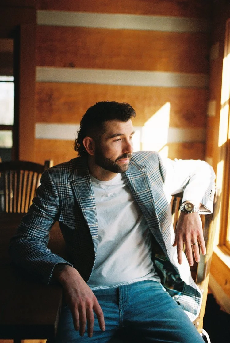 A man with dark hair and a beard sitting indoors near a window with sunlight, wearing a plaid blazer, white T-shirt, and jeans, looking thoughtfully to the side.