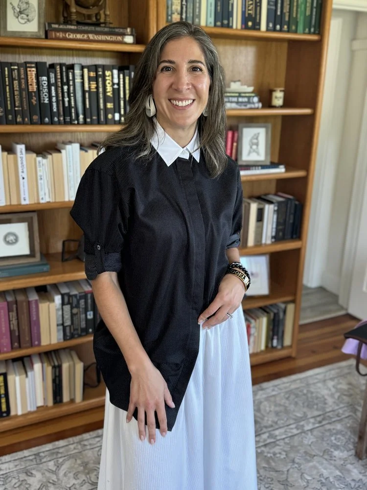 A woman with gray hair, wearing a white collared shirt, a black dress, and earrings, standing in front of a bookshelf filled with books, smiling at the camera.