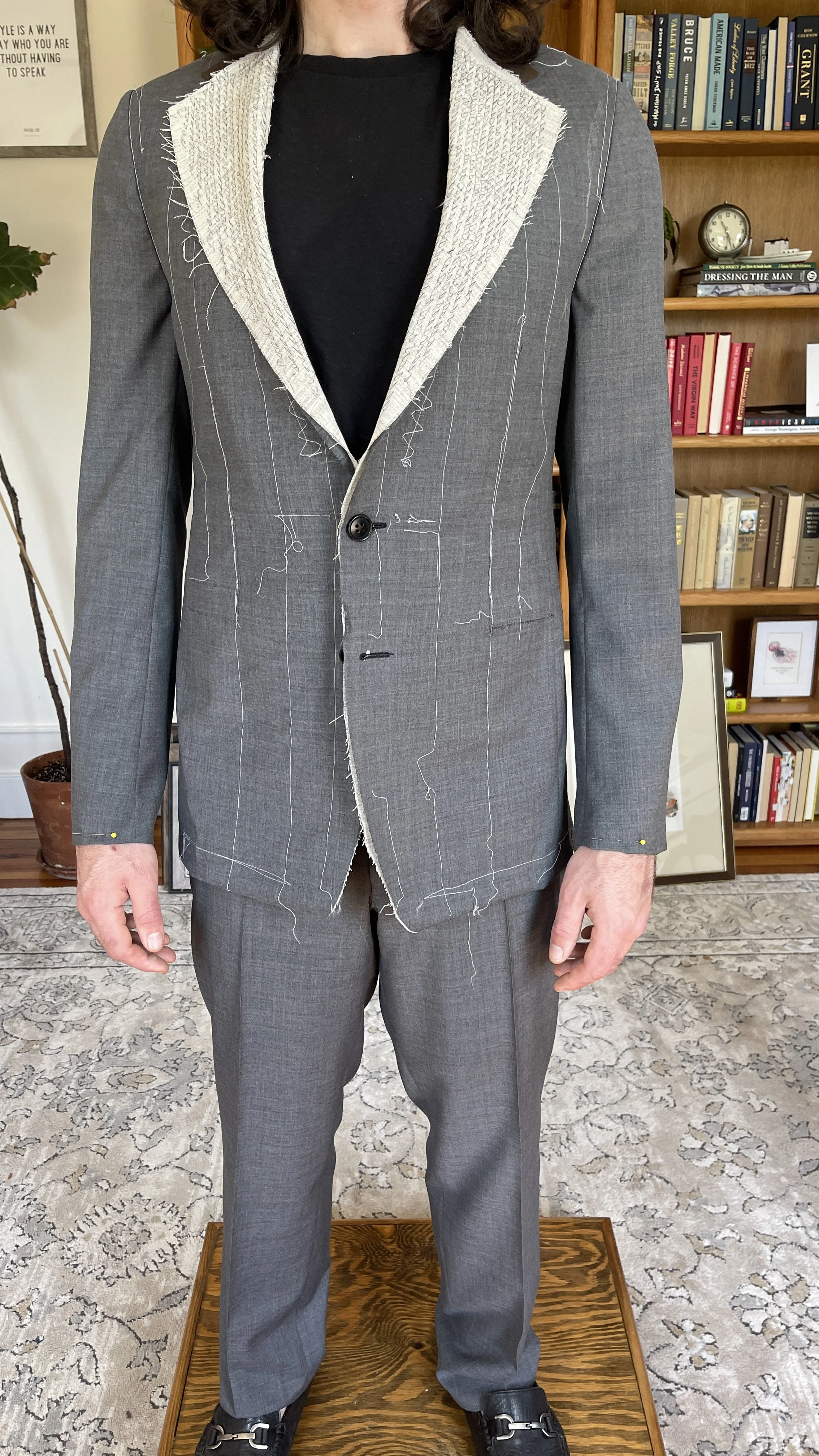 A person wearing a grey suit with visible hand-stitched chalk lines, a black shirt, and black shoes, standing on a wooden platform in a room with a bookshelf and framed pictures in the background.