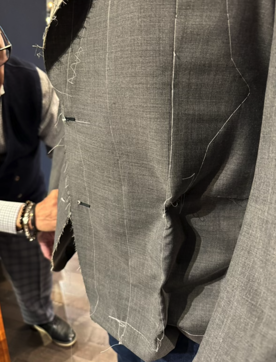 Close-up of a man wearing a gray suit jacket with visible hand-stitched tailoring details, during a fitting or tailoring process.