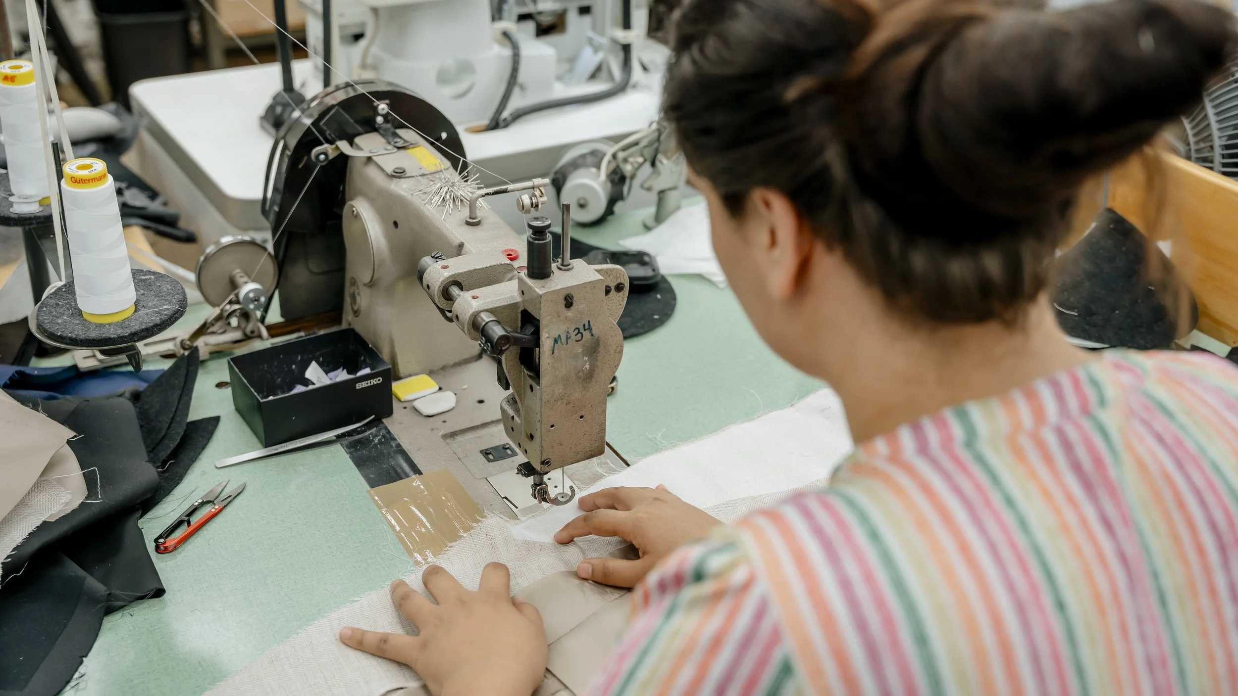 A person using a sewing machine to stitch fabric in a workshop or tailor shop, with various tools and sewing supplies on the worktable.