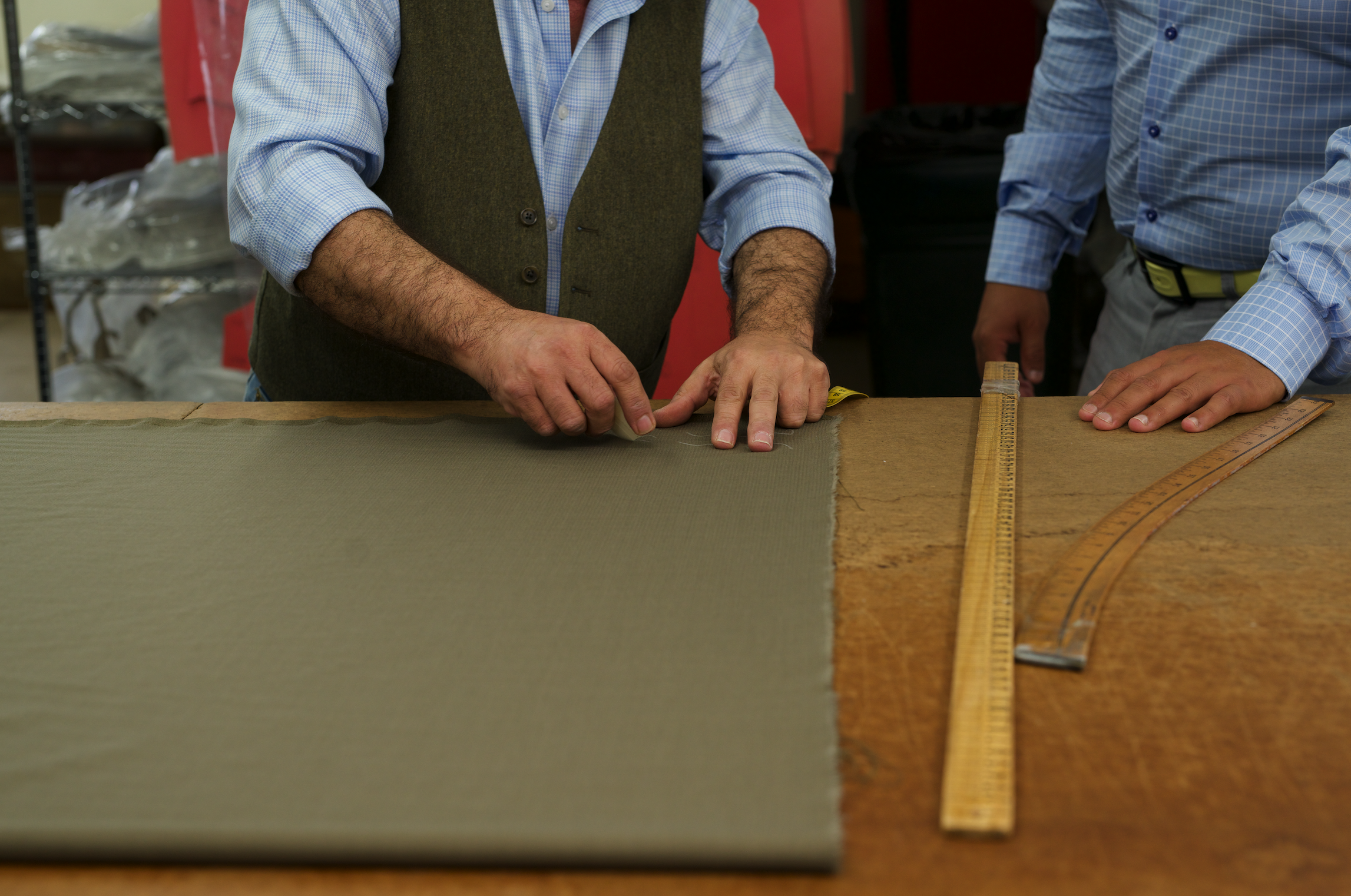 Two men working on a craft project, with one measuring or cutting fabric or material on a wooden table in a workshop or studio setting.