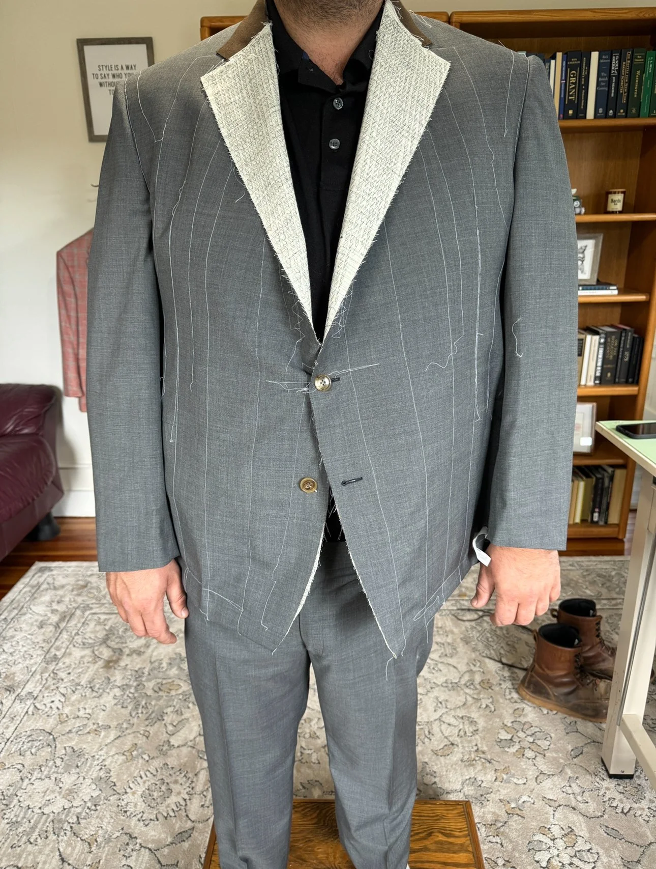 A man wearing a gray suit with a partially sewn lapel, over a black shirt, standing indoors with a bookshelf, a framed picture, and a pair of boots visible in the background.