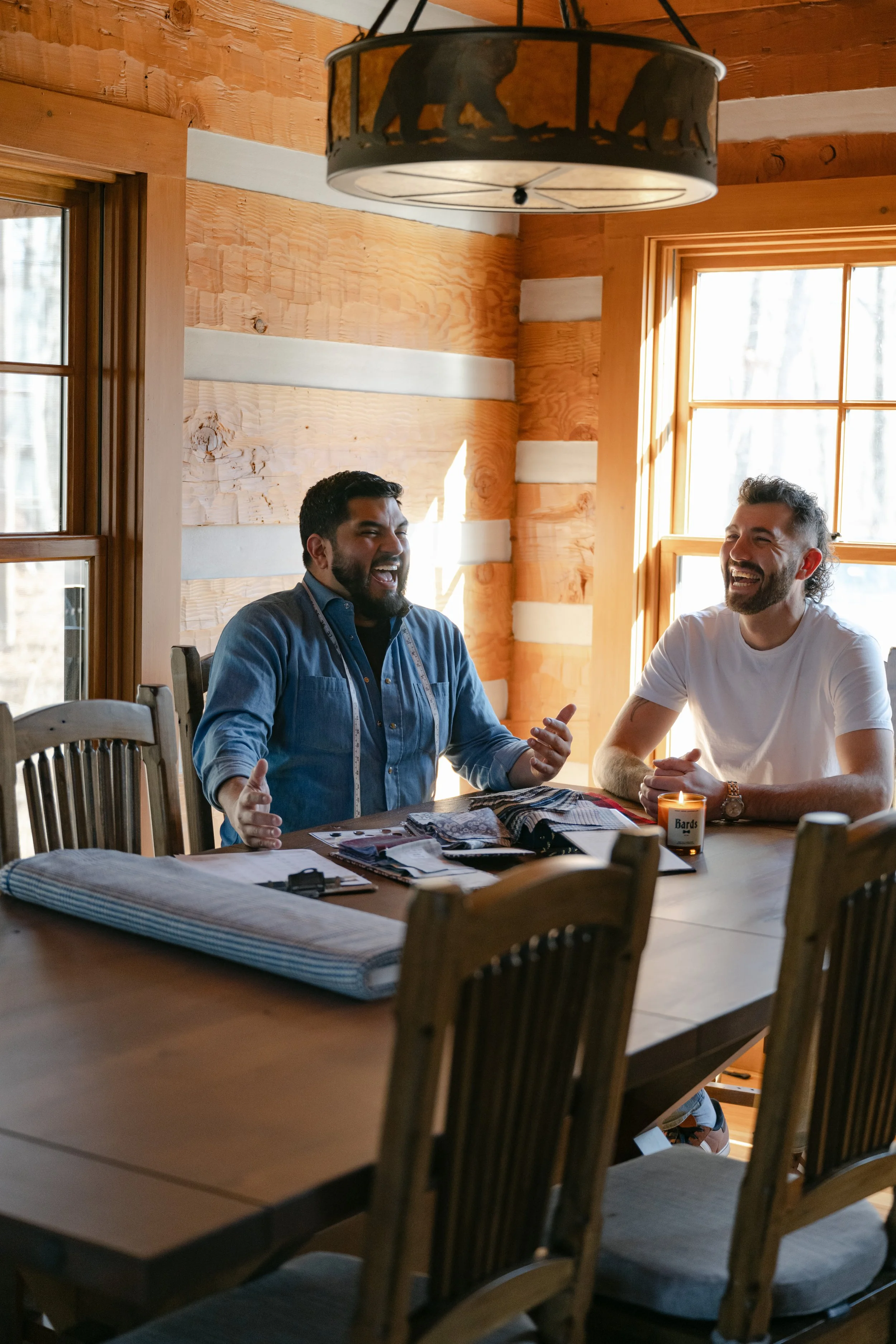 Two men sitting at a wooden dining table inside a cozy log cabin, smiling and laughing while consulting fabric swatches and design books. Natural light from large windows brightens the room, with a candle and papers scattered on the table.