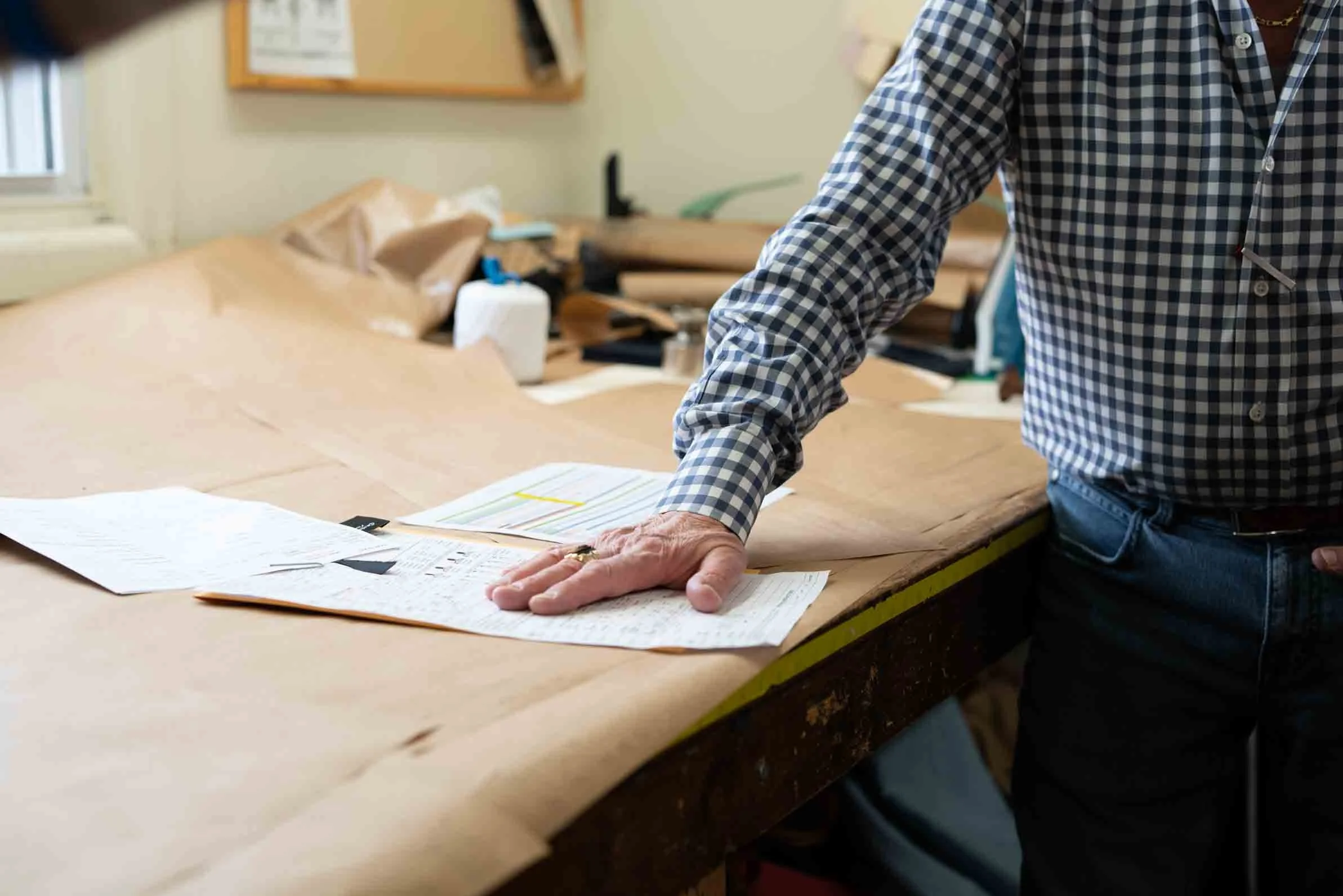 Person in checkered shirt leaning over a large workspace covered with brown paper, with papers and tools scattered on the table.