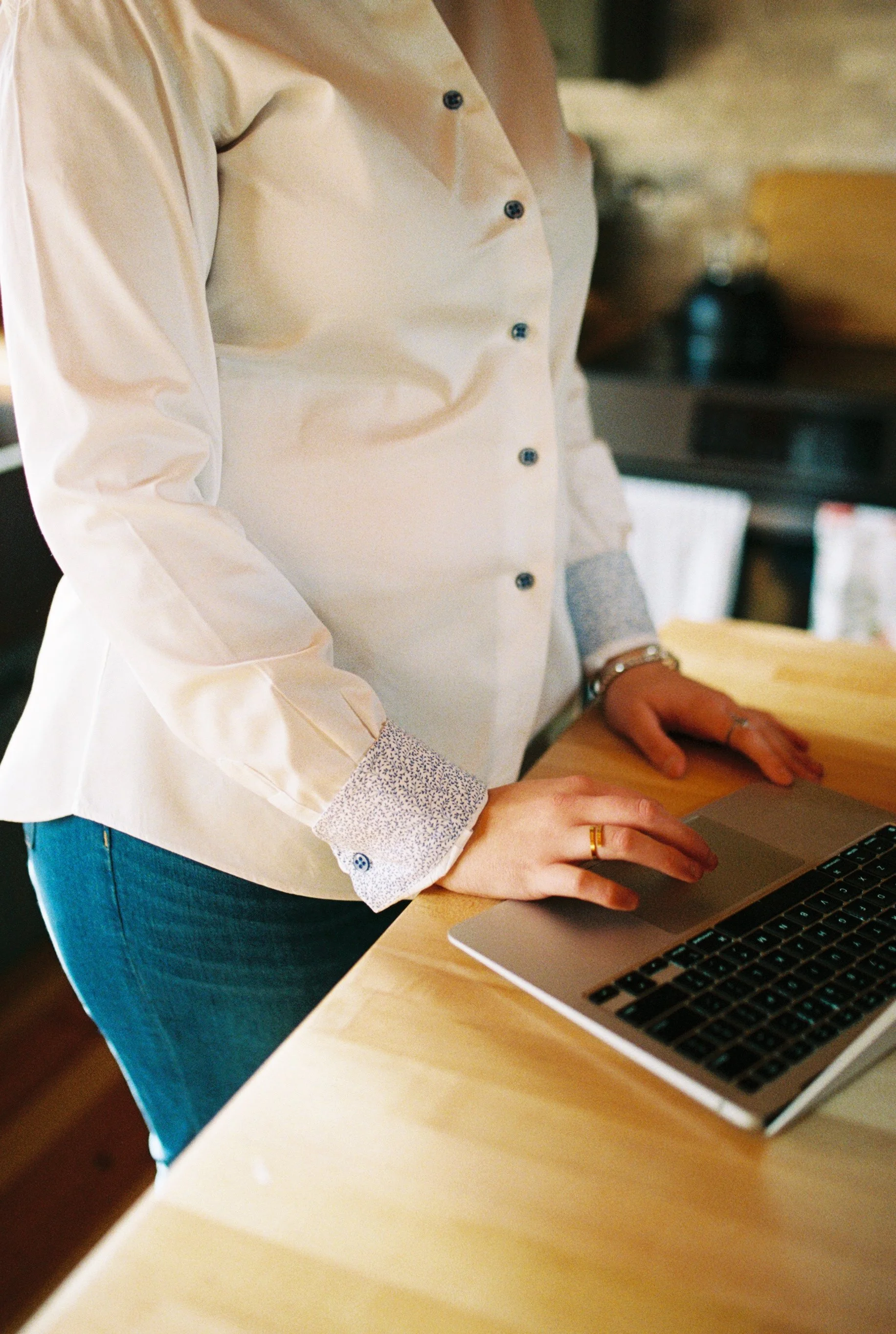 Person working on a laptop at a wooden table, wearing a cream-colored shirt with patterned cuffs and blue jeans.