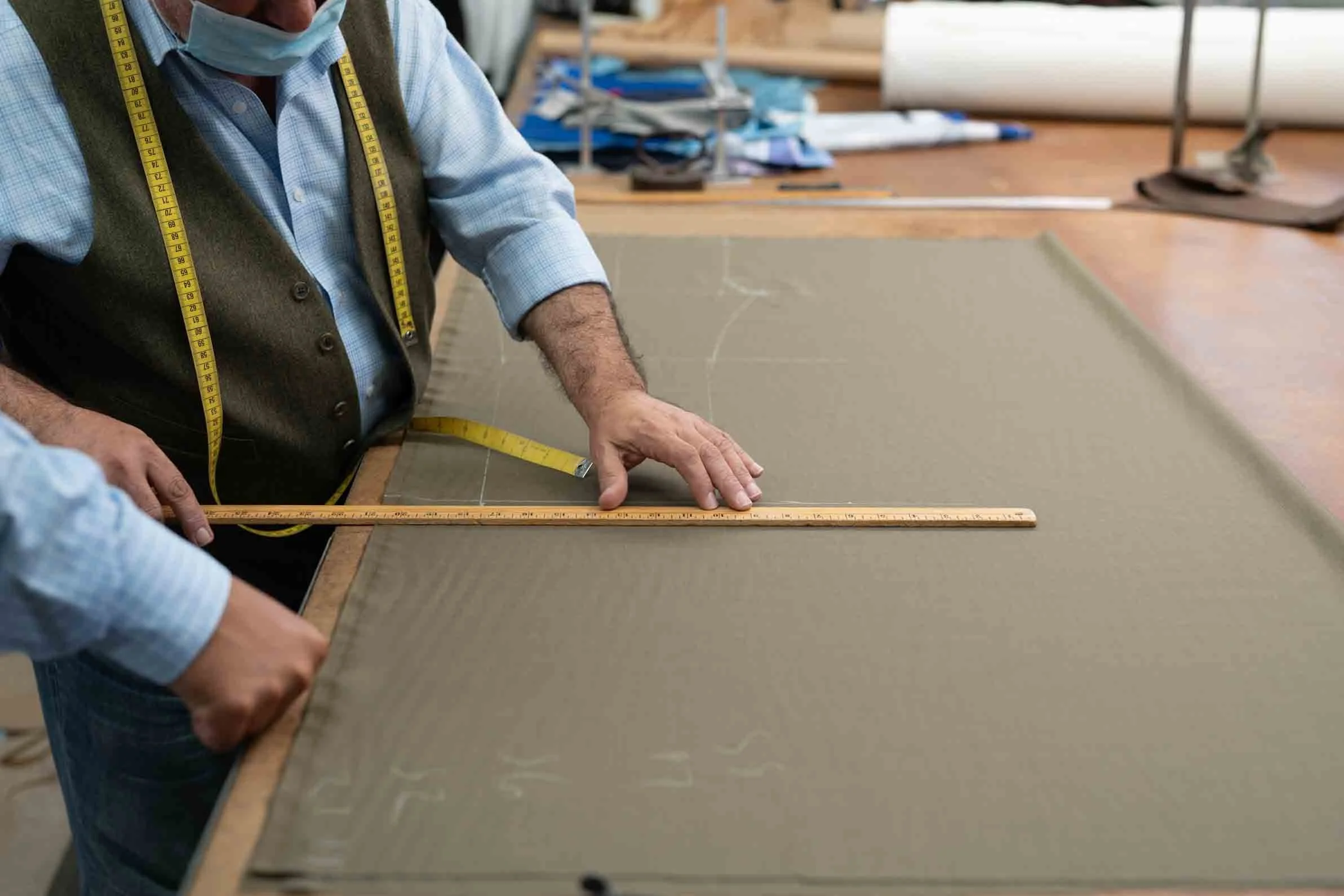 A person wearing a face mask, vest, and collared shirt measures a piece of fabric or material on a table with a ruler and a measuring tape. The workspace contains various tools and rolls of fabric or paper in the background.