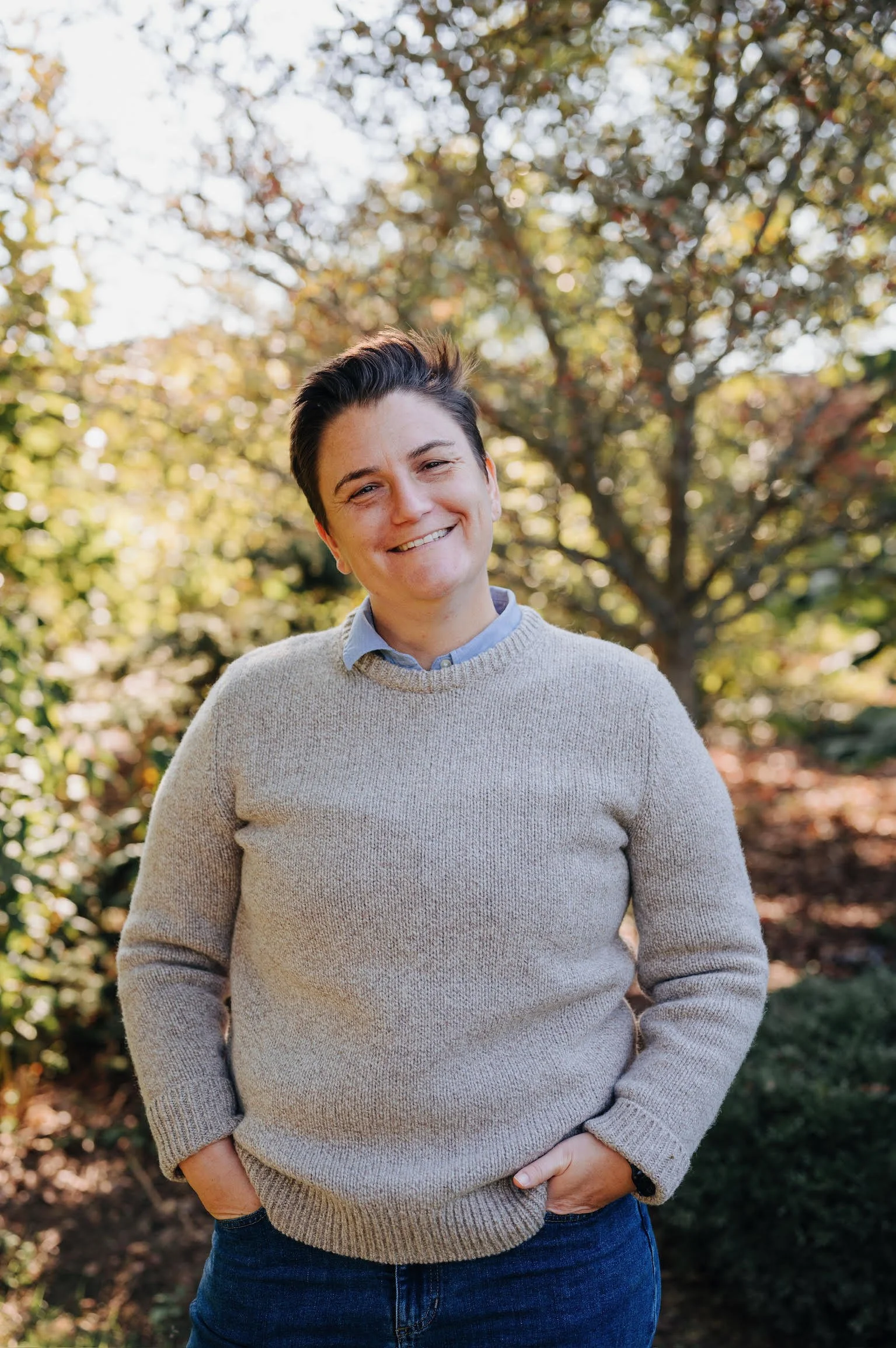 Professional headshot of Tracy Higgins where she is smiling and standing outdoors with trees and sunlight in the background.
