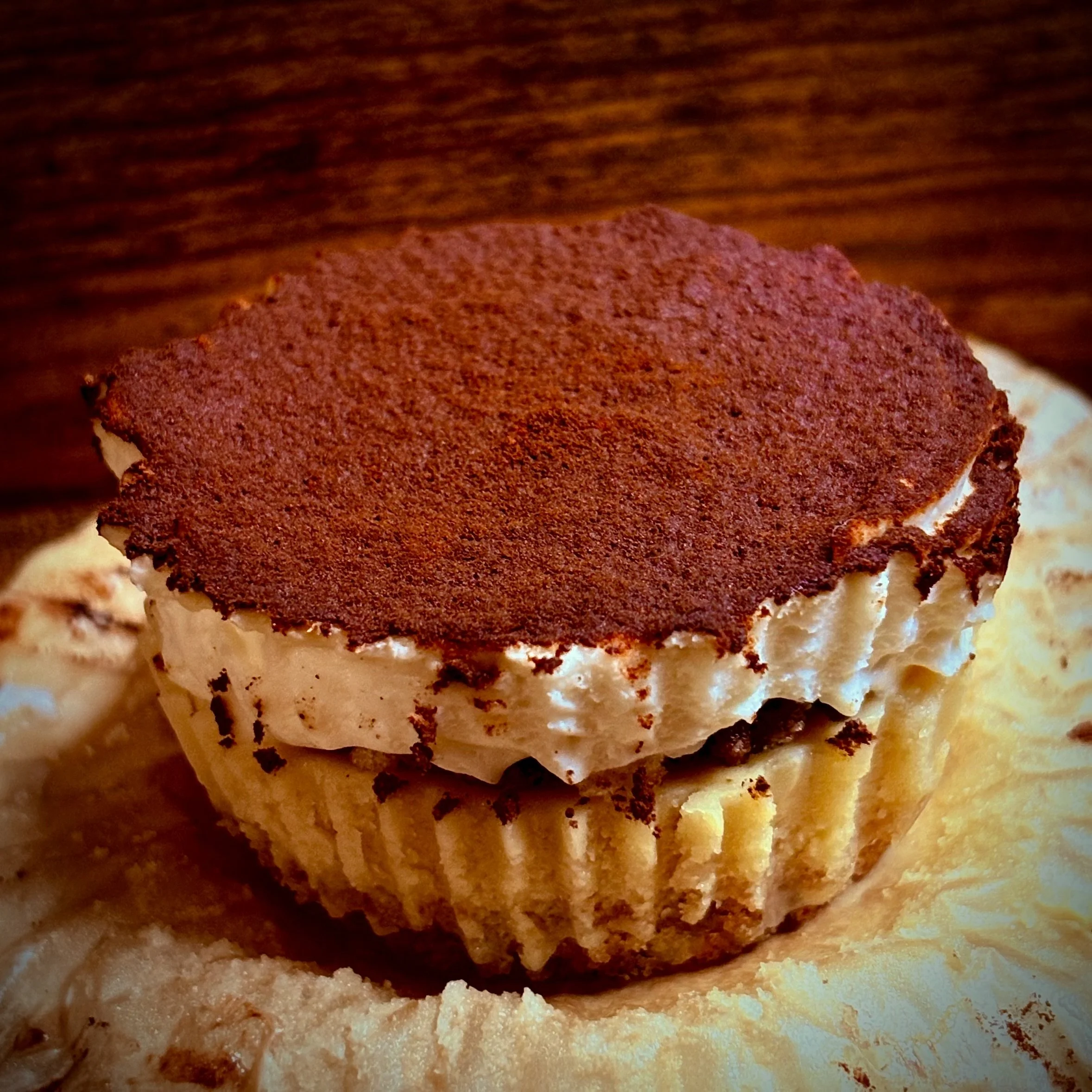 Cupcake with white frosting and a dusting of cocoa powder on top, resting on a wooden surface.