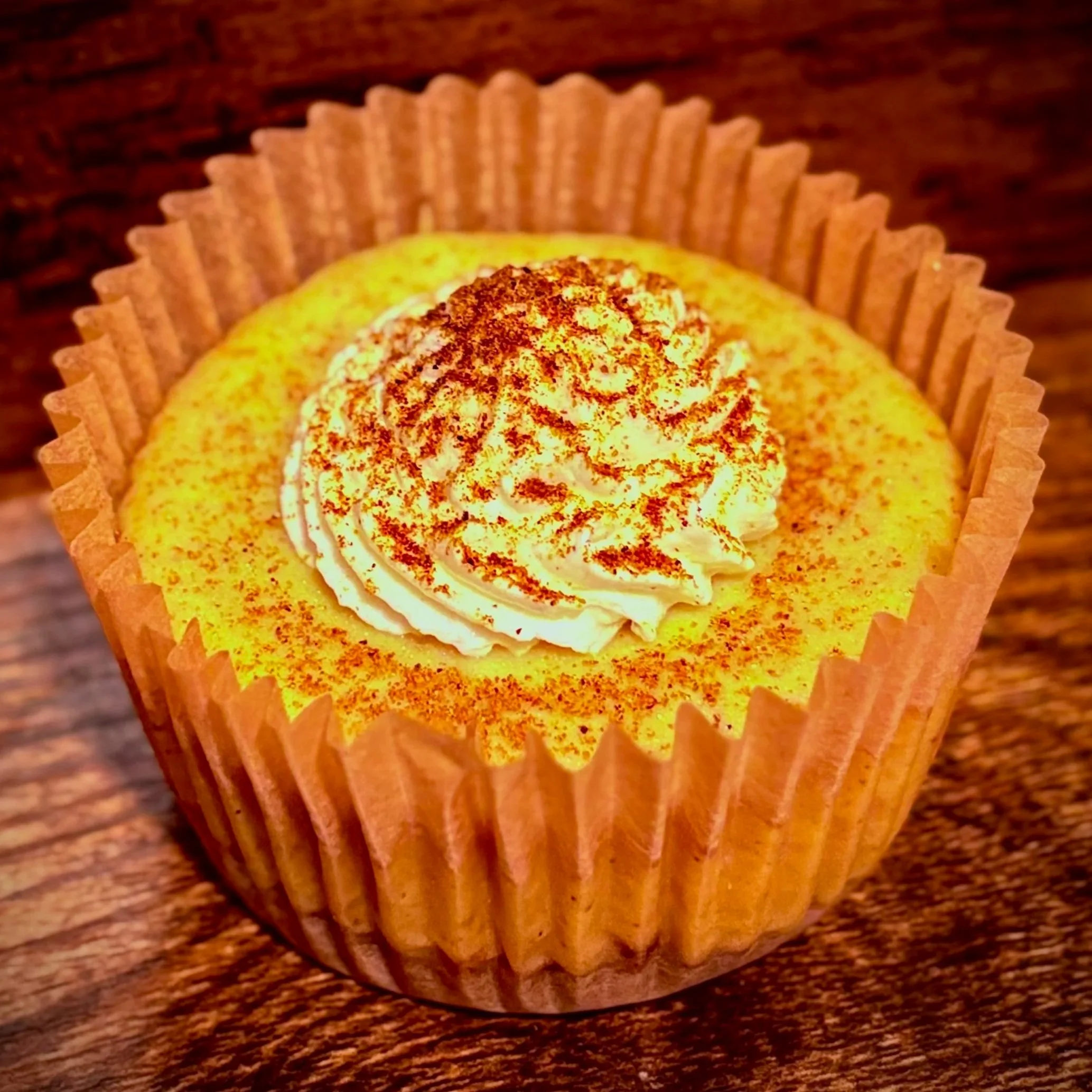 A cupcake with yellow frosting, topped with whipped cream and sprinkled with cinnamon, in a paper cupcake liner on a wooden surface.