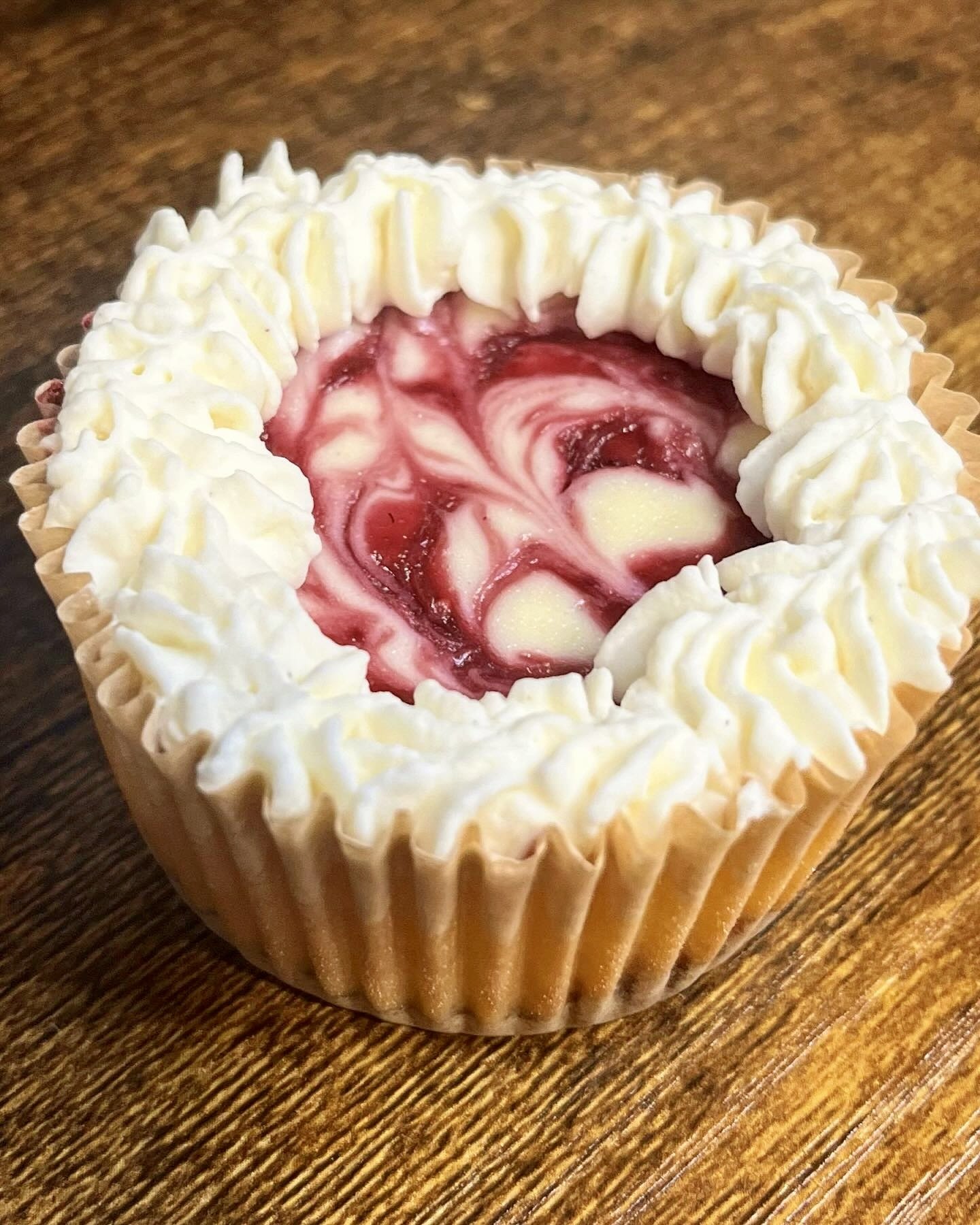 A cupcake with white frosting and a red and white swirled fruit topping, in a paper liner on a wooden surface.
