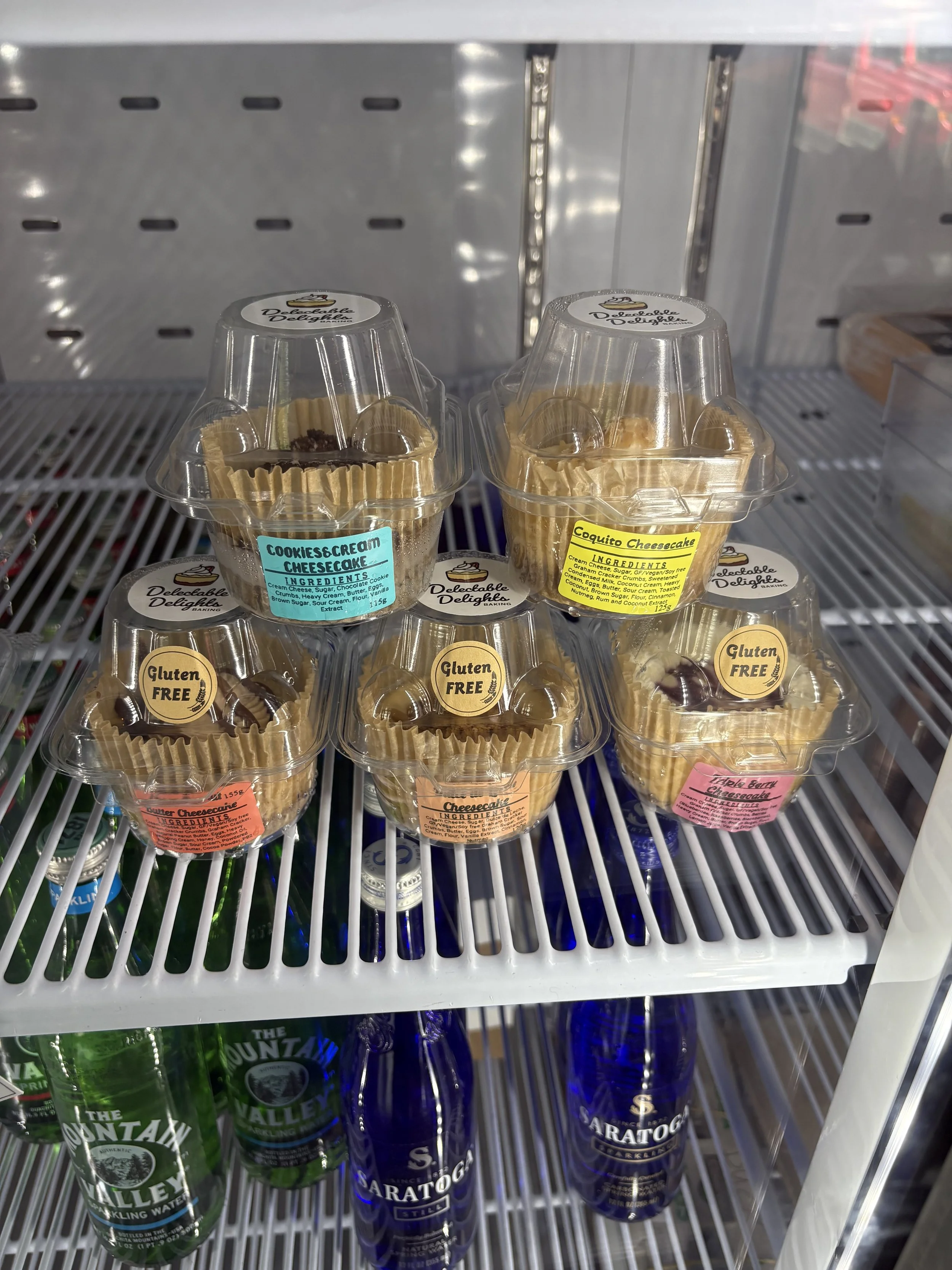 Three cupcakes with labels reading 'Cookies & Cream', 'Coquito Cheesecake', and 'Triple Berry Cheesecake' are displayed in a refrigerated case, with bottles of water and soda below.