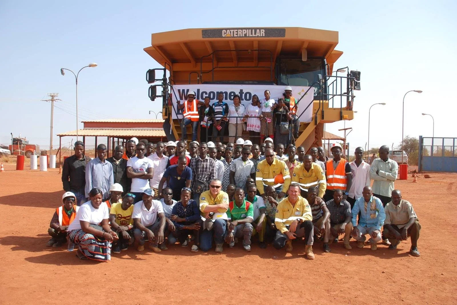 Group of people standing and sitting in front of a large Caterpillar construction vehicle. The group includes men and women, some wearing safety vests and helmets. The background shows a clear sky, some small buildings, and streetlights, all on a reddish dirt ground.